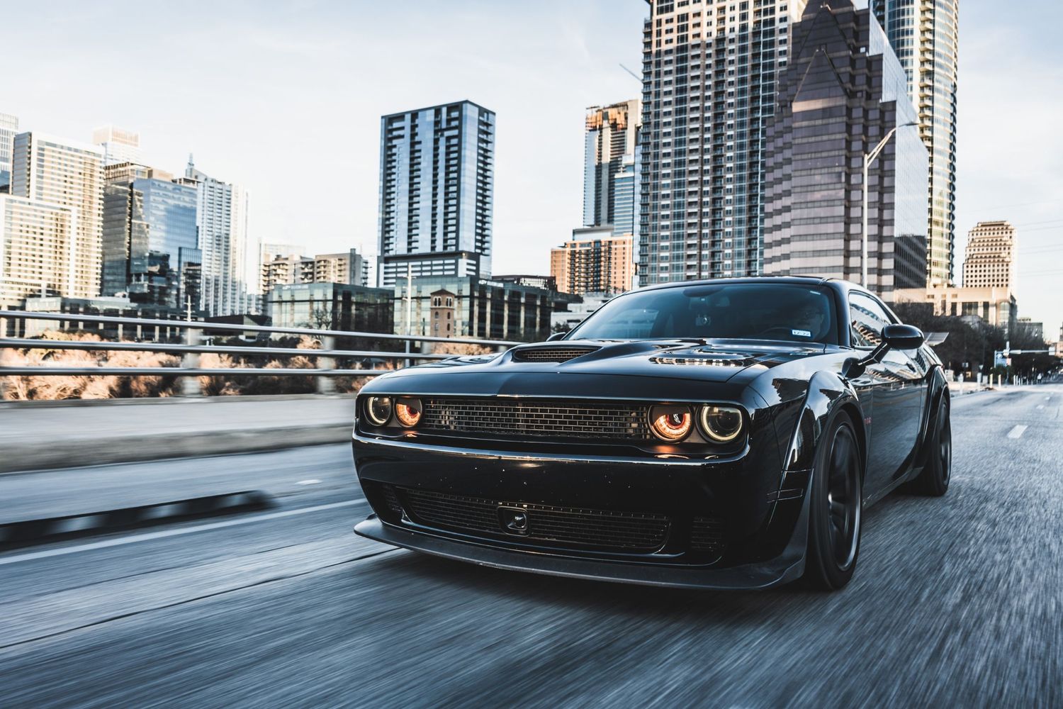 Matte black Dodge Challenger speeds past modern city architecture with glowing orange headlights.