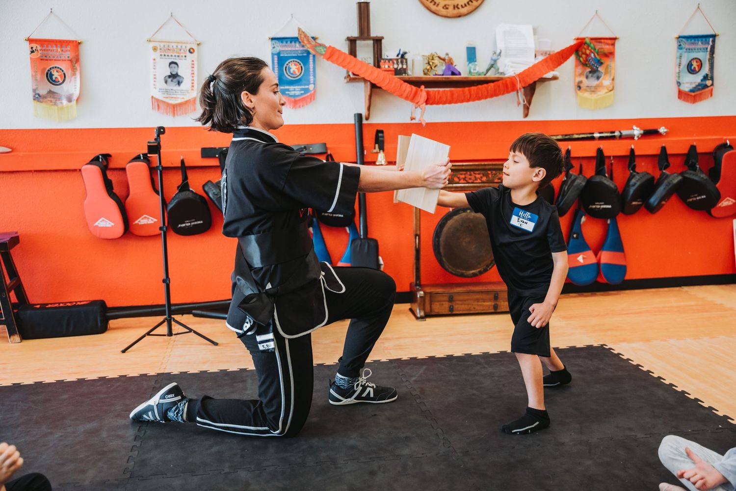 Instructor demonstrates martial arts technique to young student in orange and black training room.