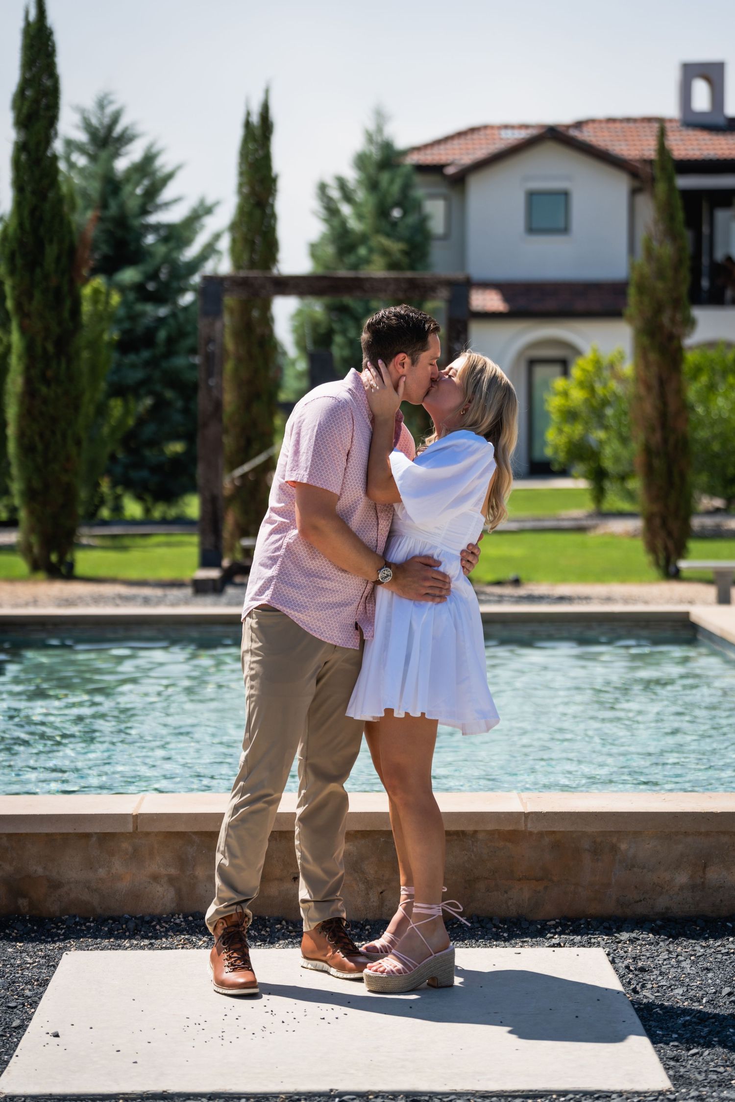 A tender embrace between a couple by a pool with cypress trees and Spanish-style architecture in background.