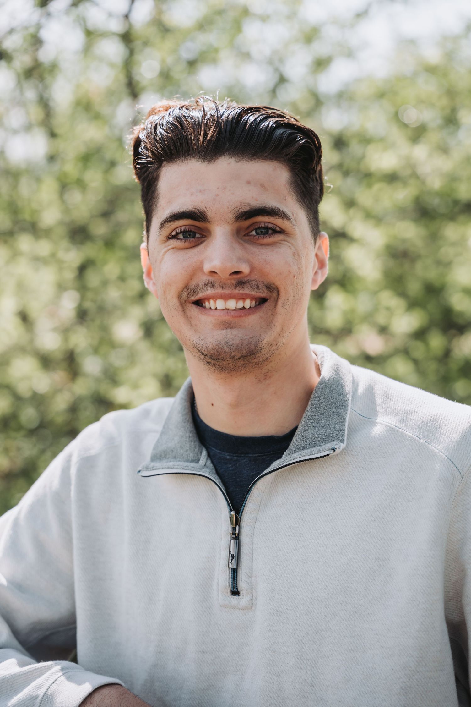 Portrait of someone in a gray quarter-zip sweater smiling outdoors with trees in background.