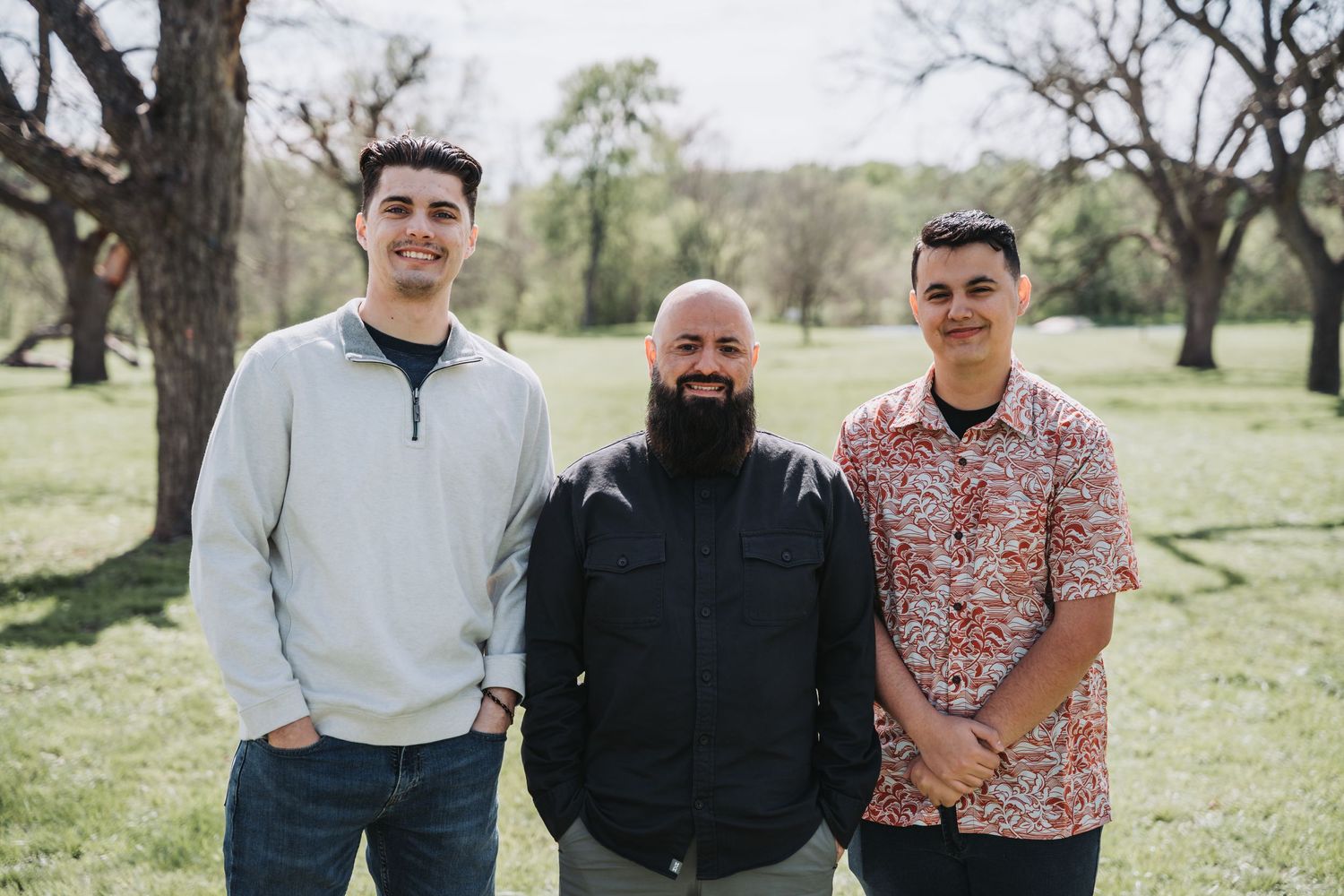Three people posing together for a casual outdoor photo with trees in the background.