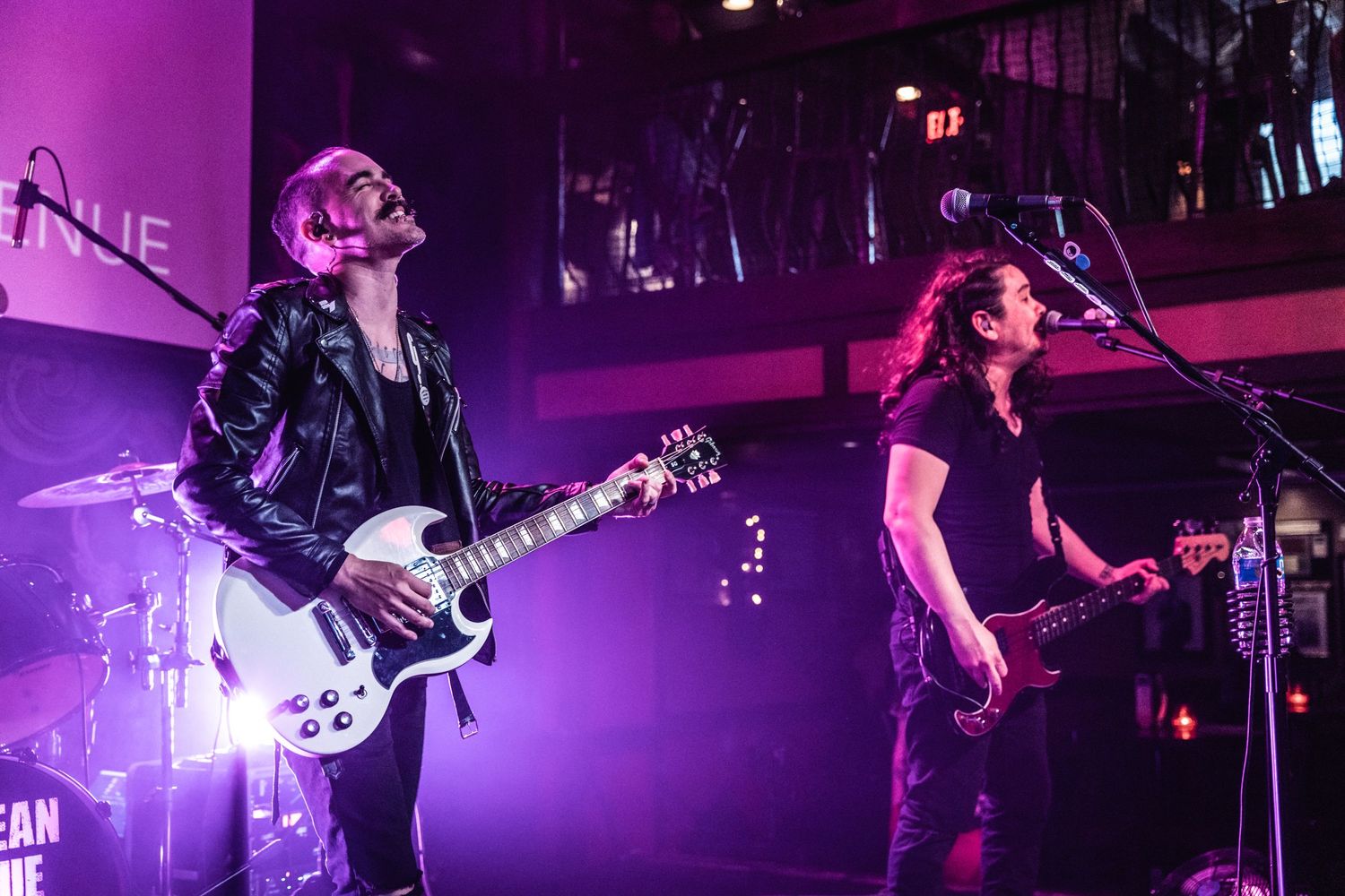 Two musicians performing on stage bathed in purple concert lighting.