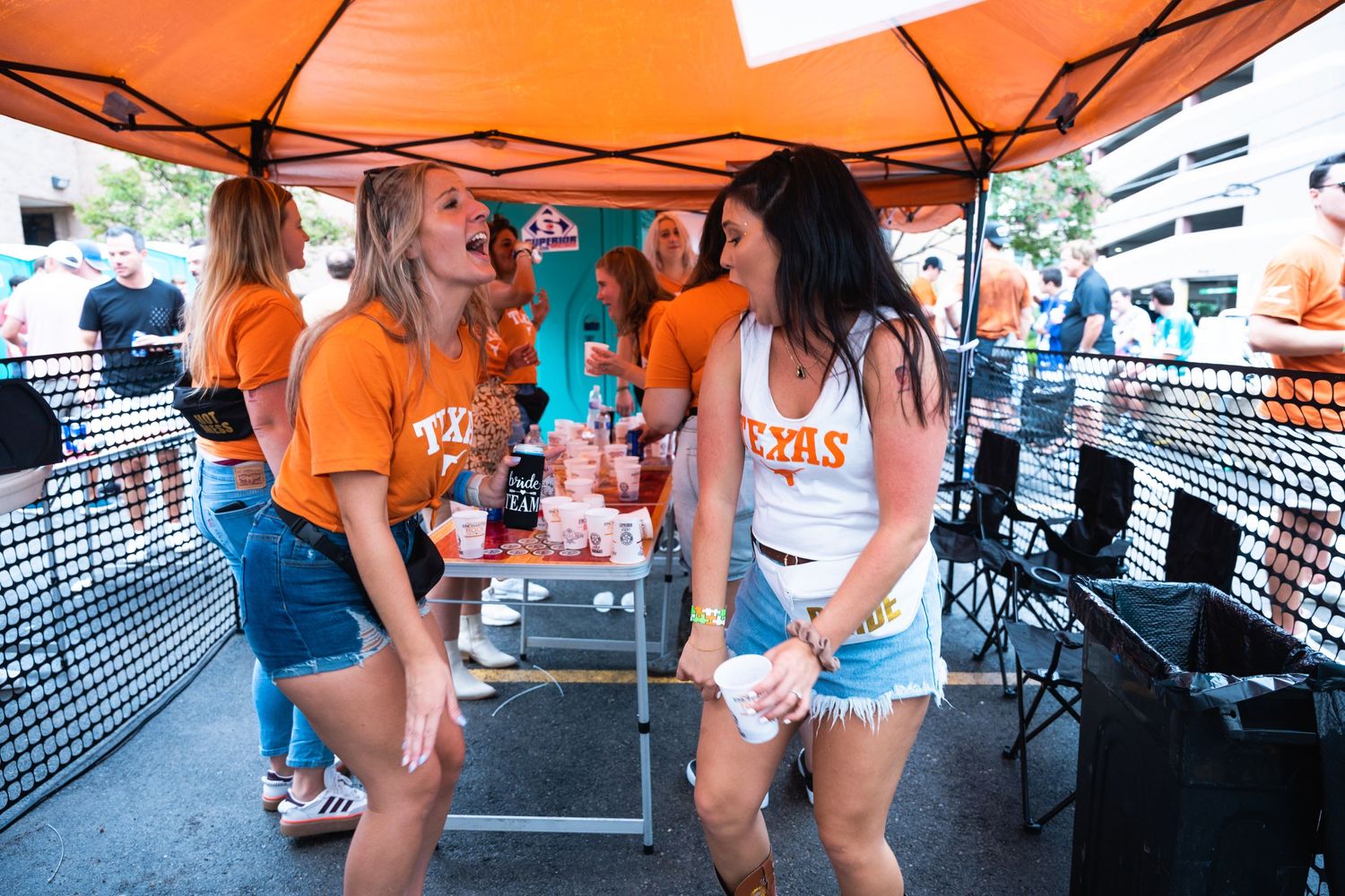 Friends in orange Texas shirts and denim shorts socialize at an outdoor party under an orange canopy.