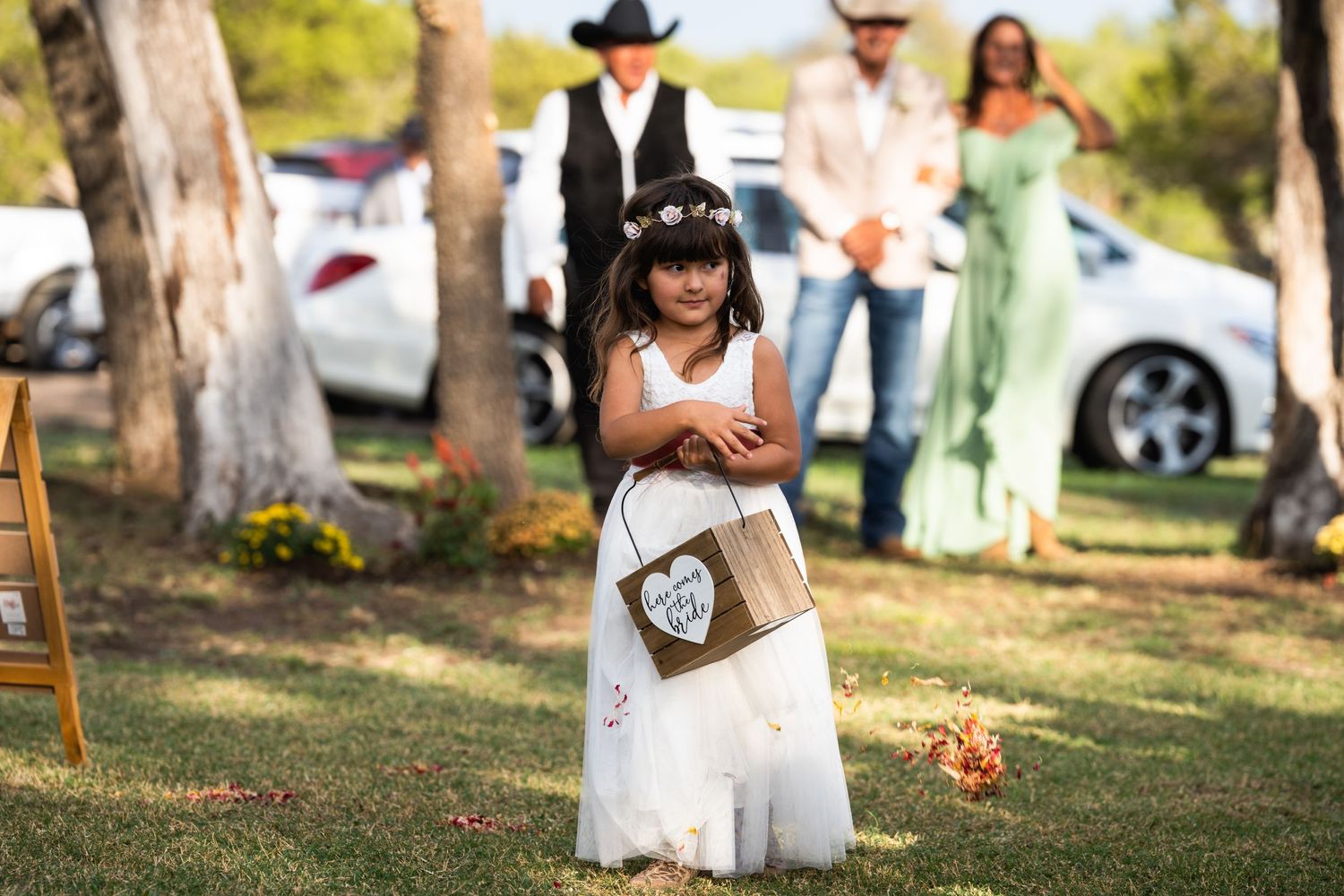 A white lace-clad flower girl holds a wooden sign at an outdoor wedding ceremony.