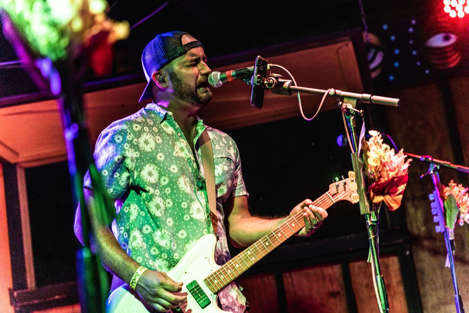 A performer plays guitar and sings into a microphone while wearing a green patterned shirt.