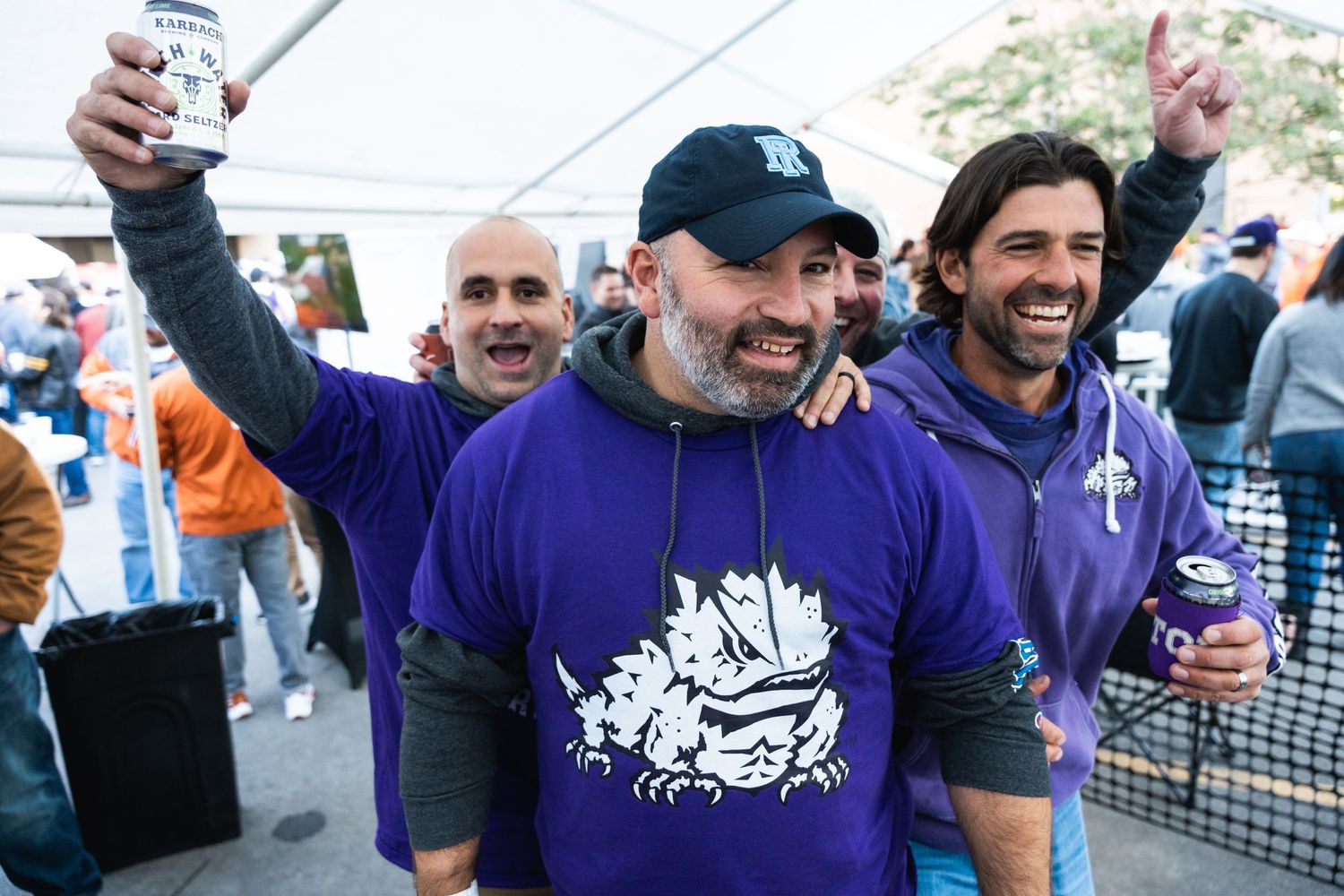 Three friends in purple team gear celebrate with drinks at an outdoor gathering.