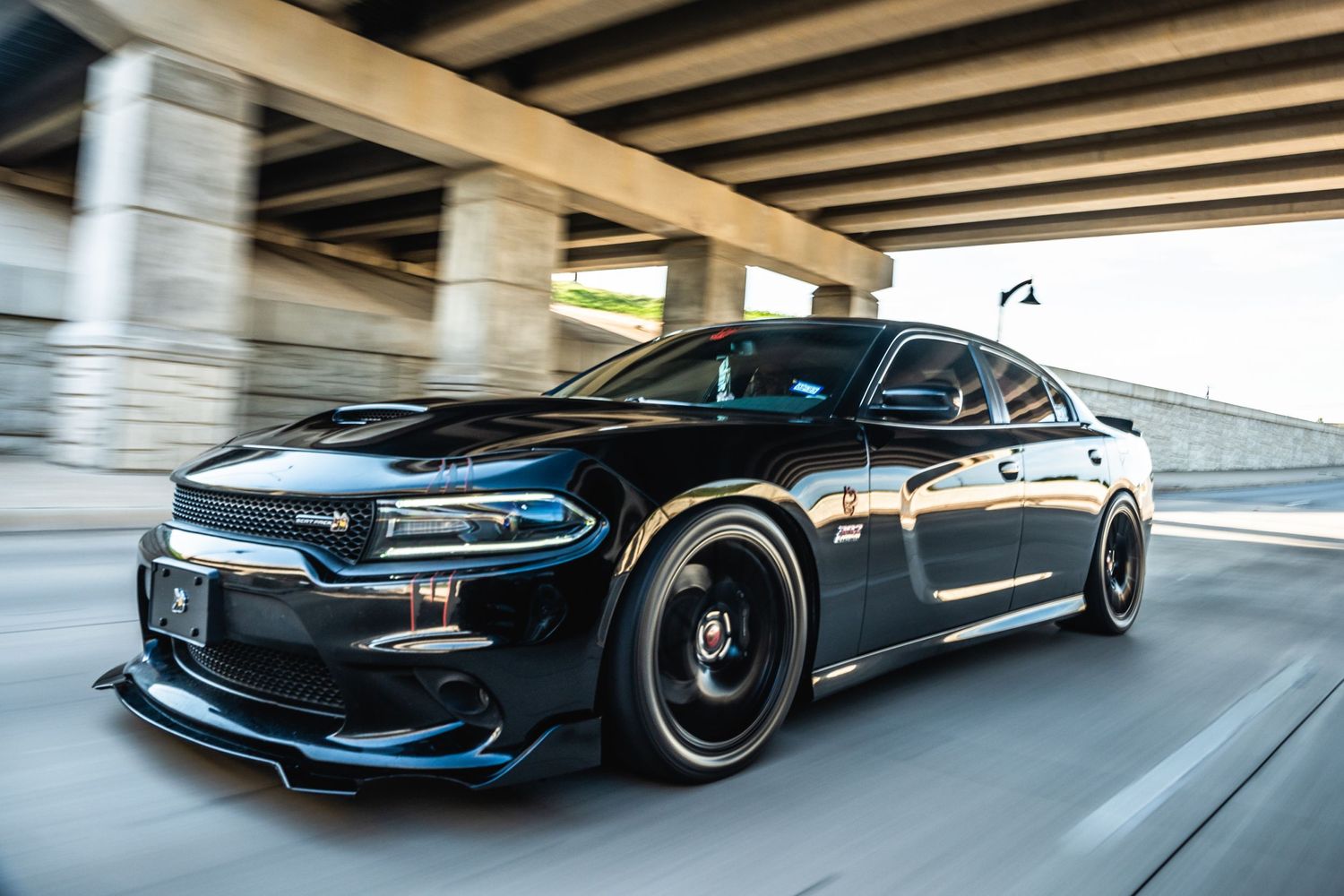 Modified black Dodge Charger racing under highway overpass.