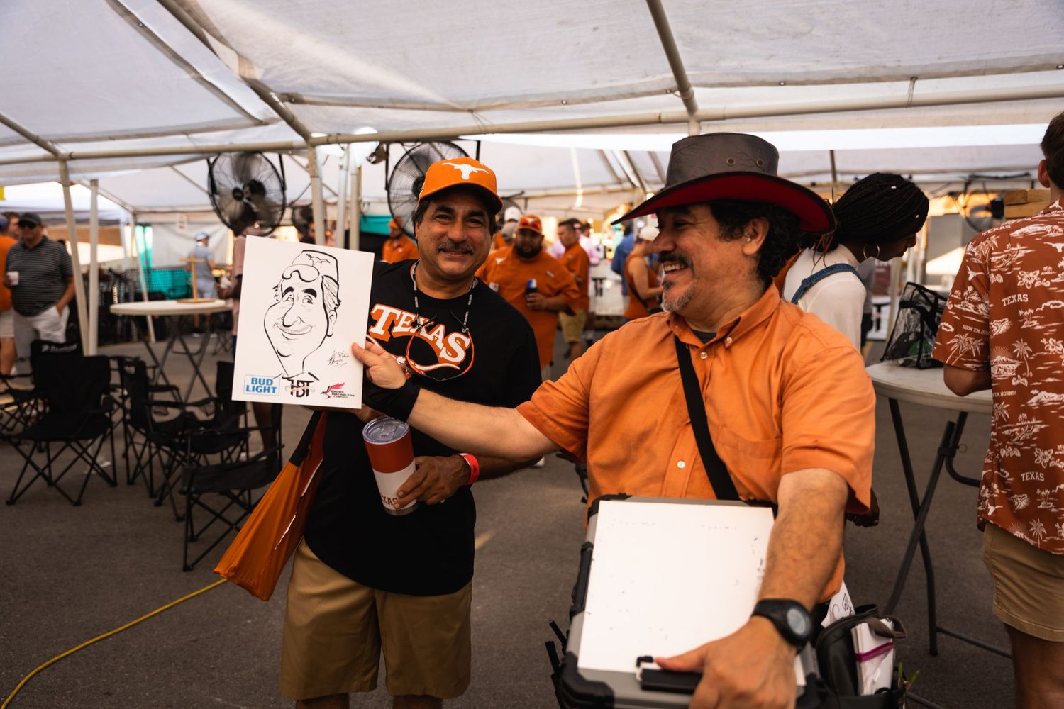 Two people share a laugh while looking at caricature drawings under a white tent.