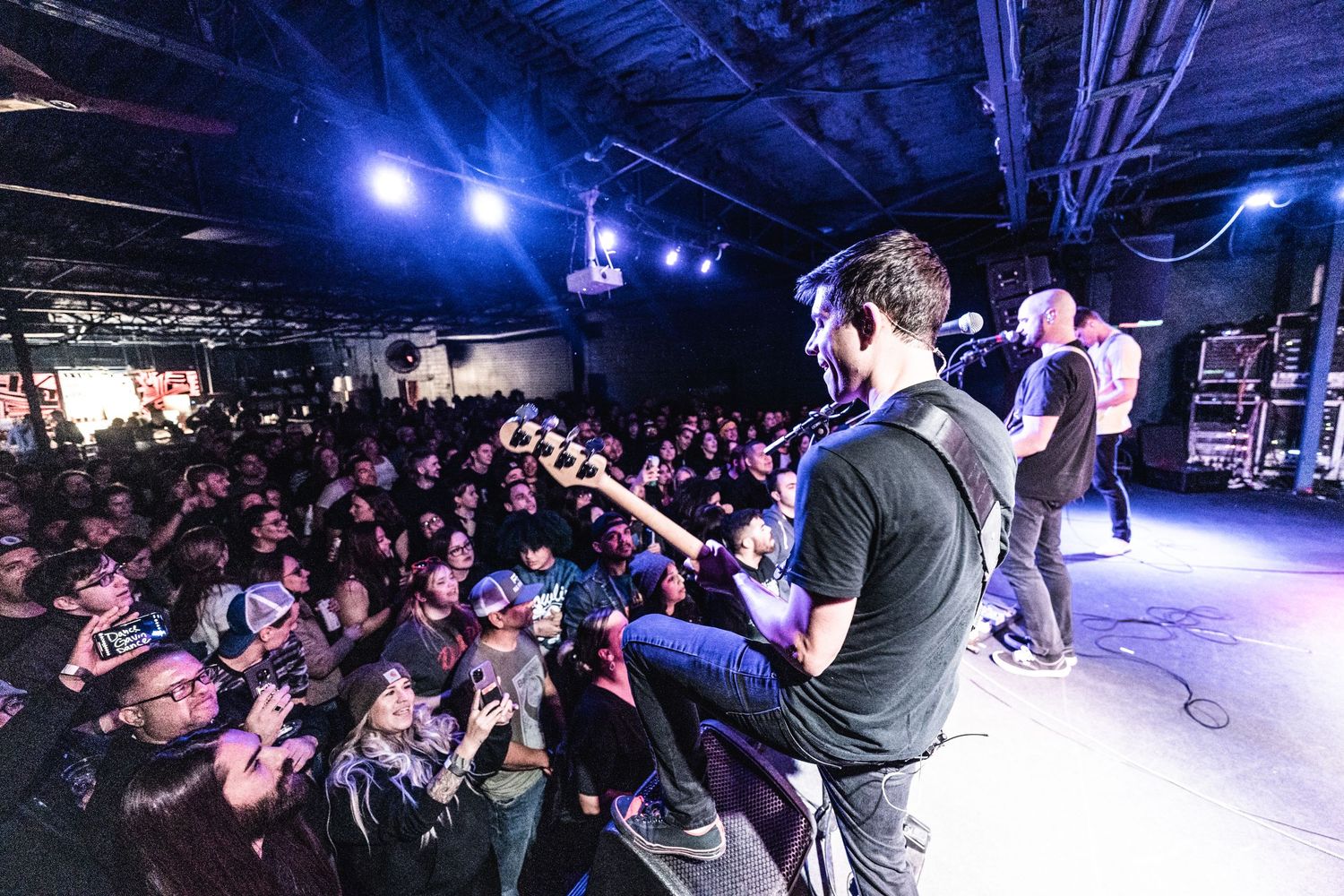 Musicians perform on stage while crowd reaches up from pit area under blue lighting.