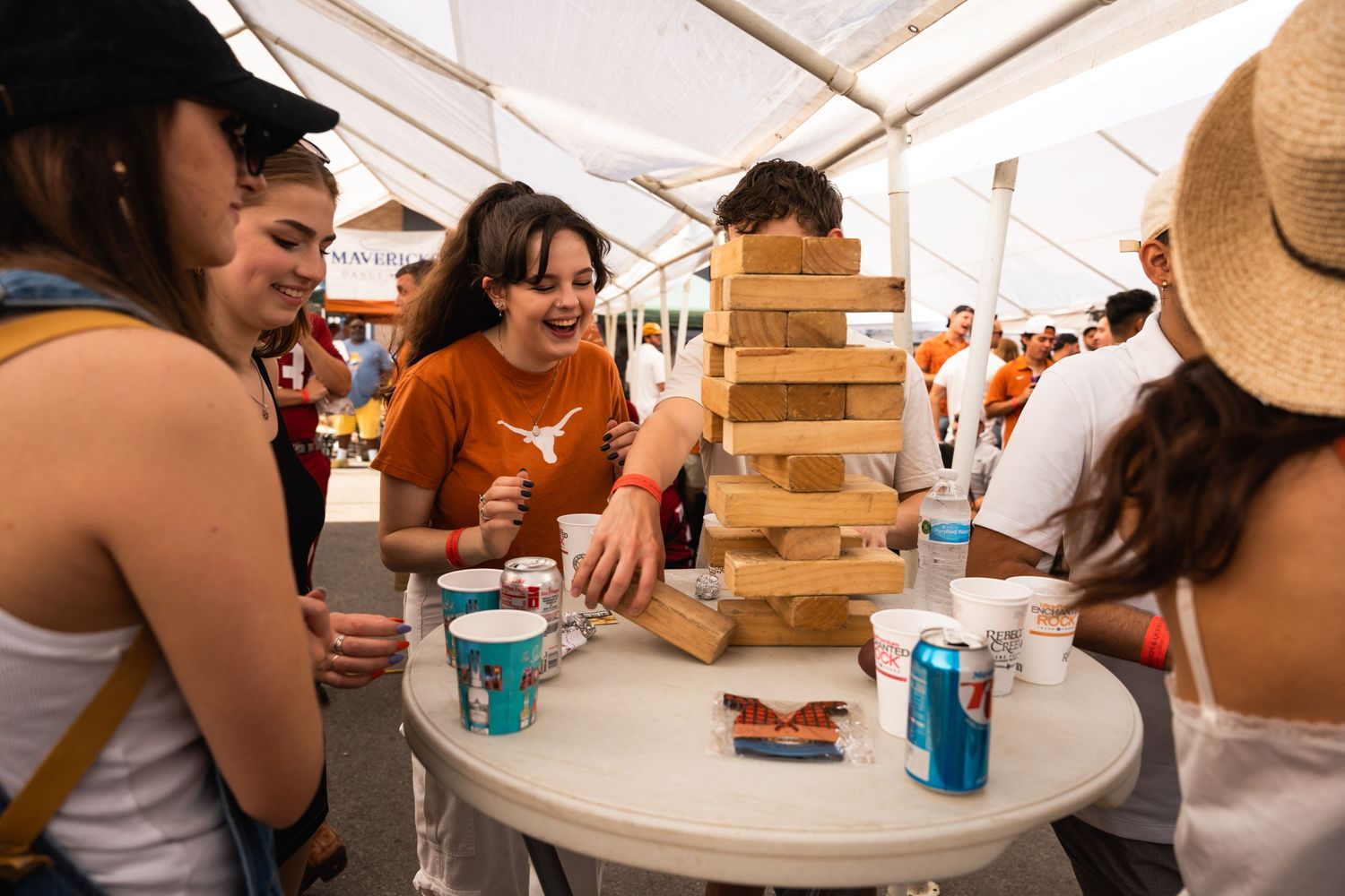 Friends gathered around a table playing Jenga under a tent at an outdoor event.