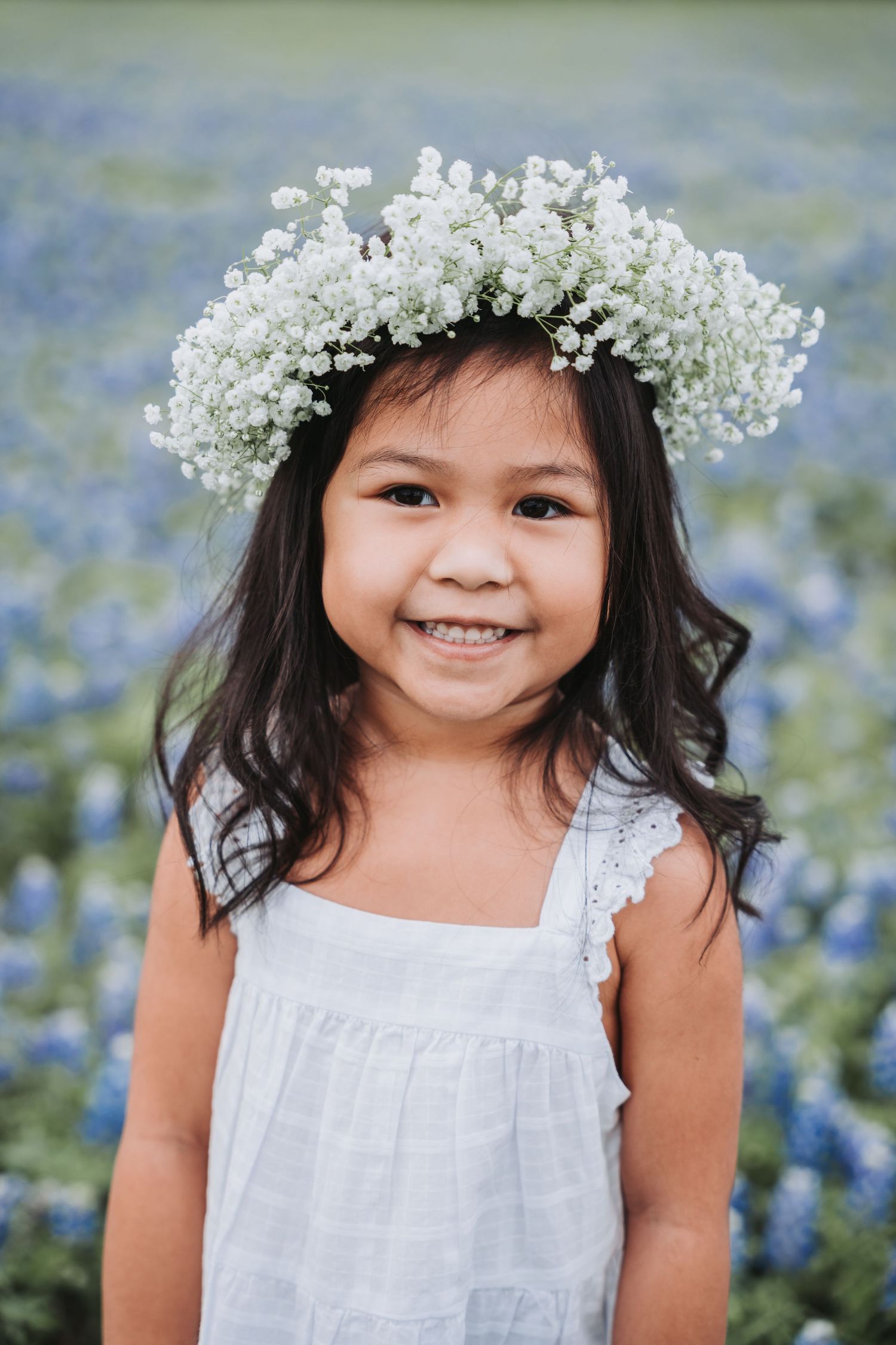 Figure in white dress wearing delicate baby's breath crown smiles in bluebonnet field.