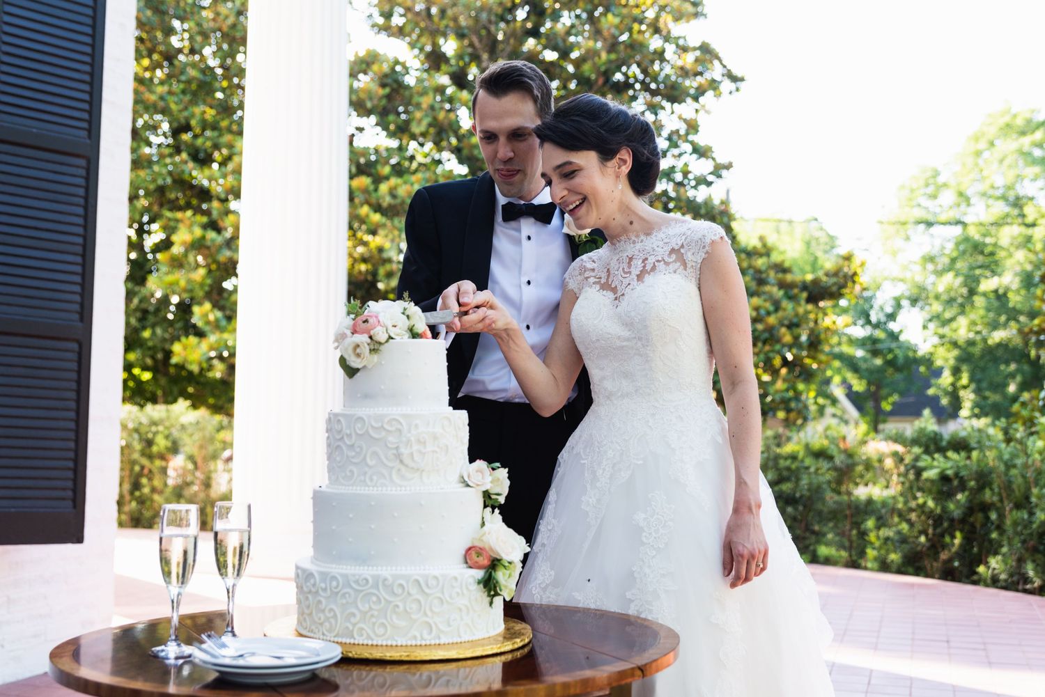 Newlyweds cutting their elegant white wedding cake together on a porch.