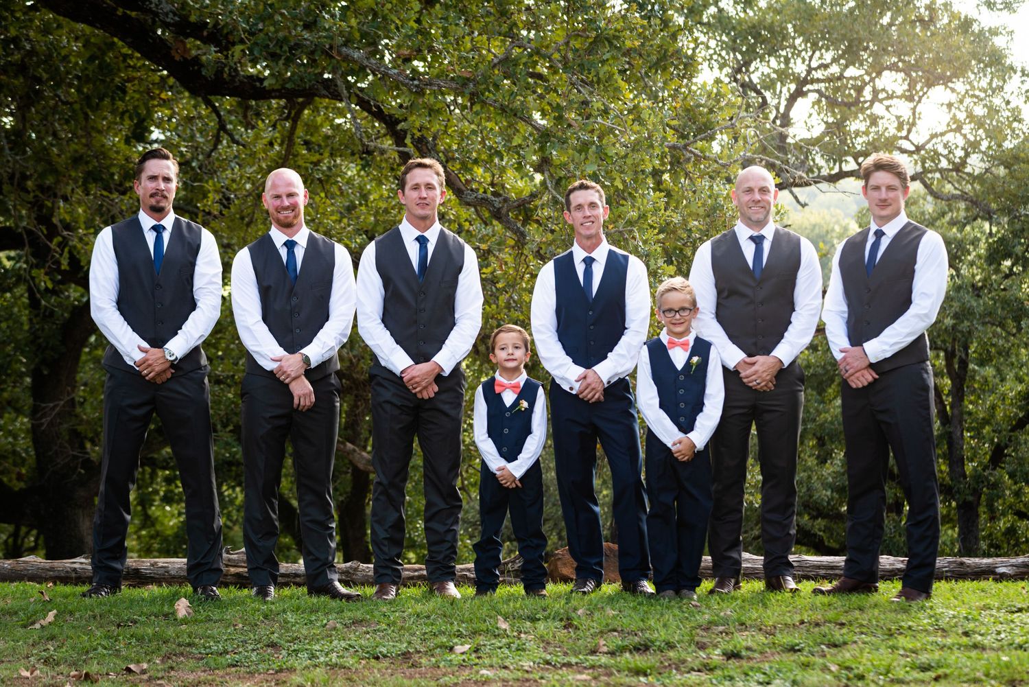 Wedding party groomsmen in matching navy vests lined up outdoors with trees in background.