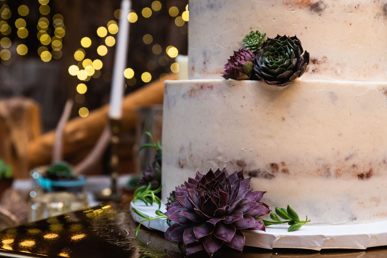 Close-up of a white wedding cake with purple succulents against bokeh lights.