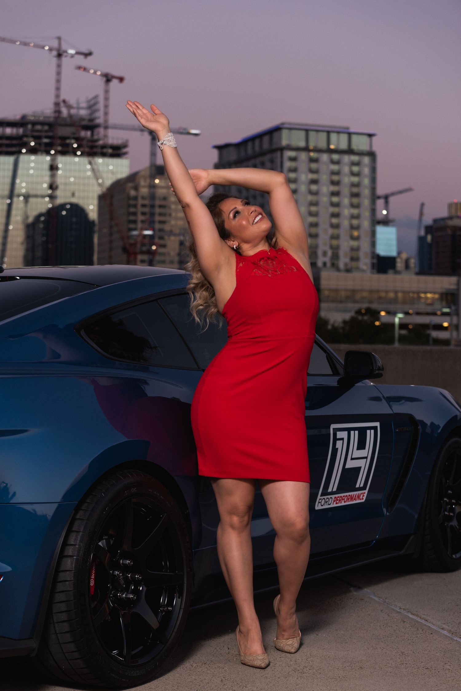 Dynamic pose against blue sports car with city buildings in background at dusk.