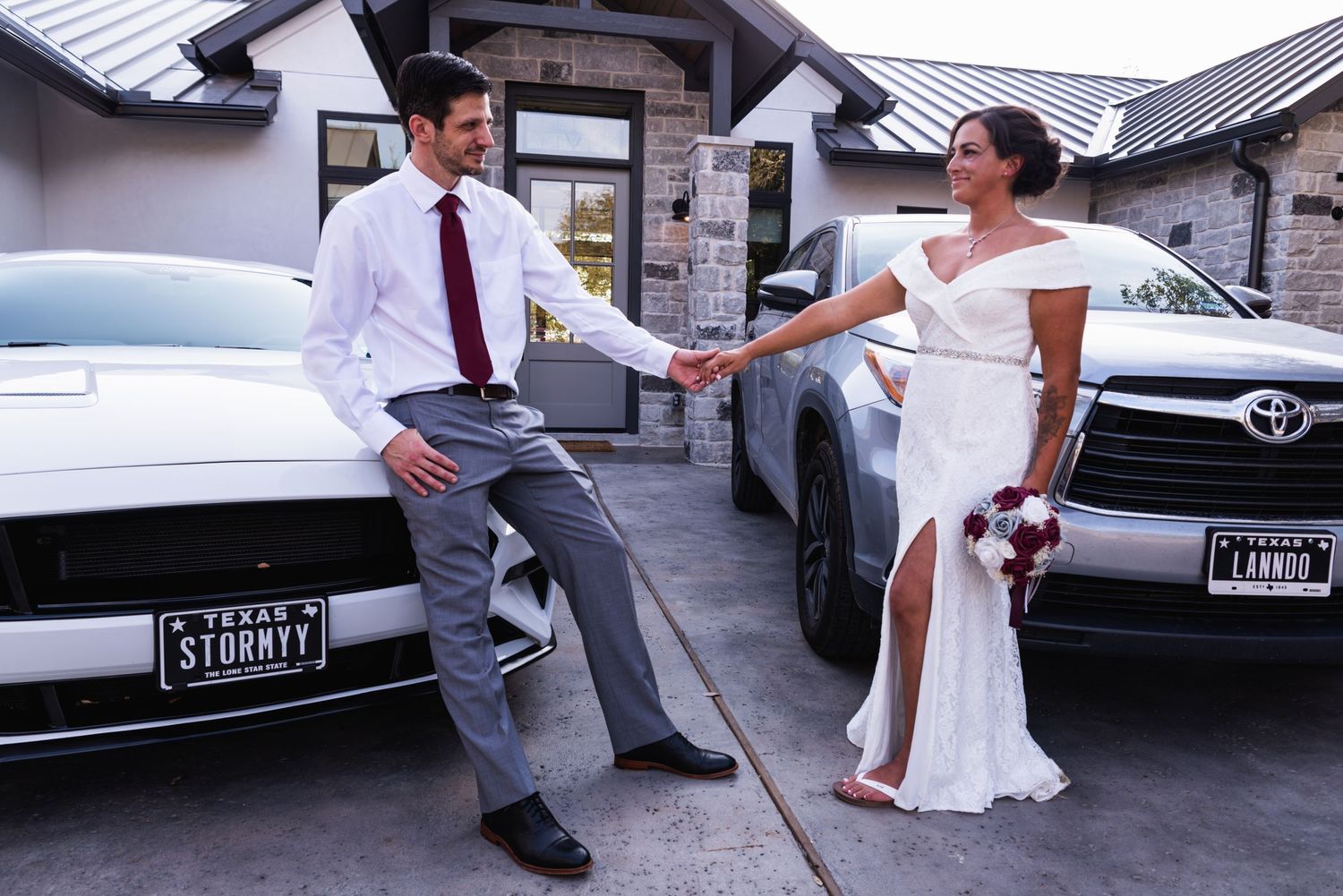 A joyful wedding couple dances playfully in a parking lot between parked cars.