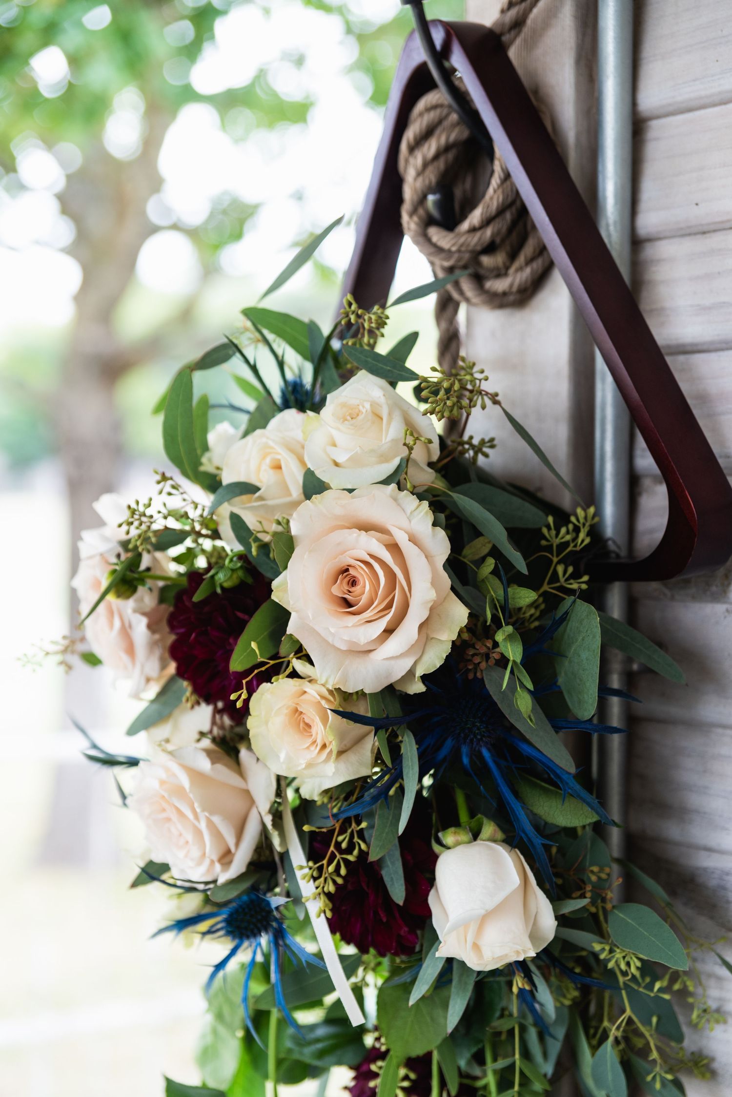 Elegant white rose and greenery wedding bouquet hanging from rustic rope against white wall.