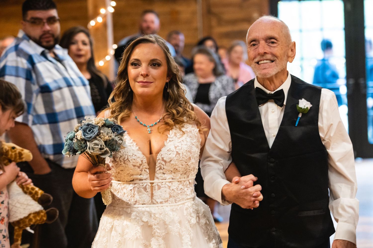 A bride in a lace wedding dress walks down the aisle holding a bouquet of flowers.