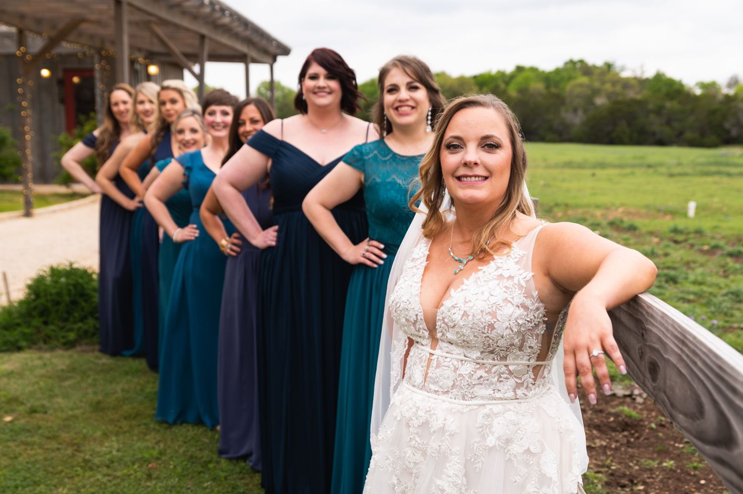 Wedding party in coordinating blue dresses posing casually along rustic railing.