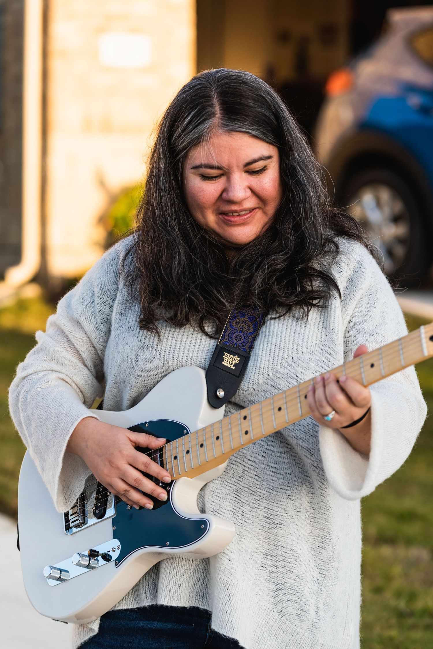 A musician plays a light blue electric guitar while wearing a cozy white sweater outdoors.