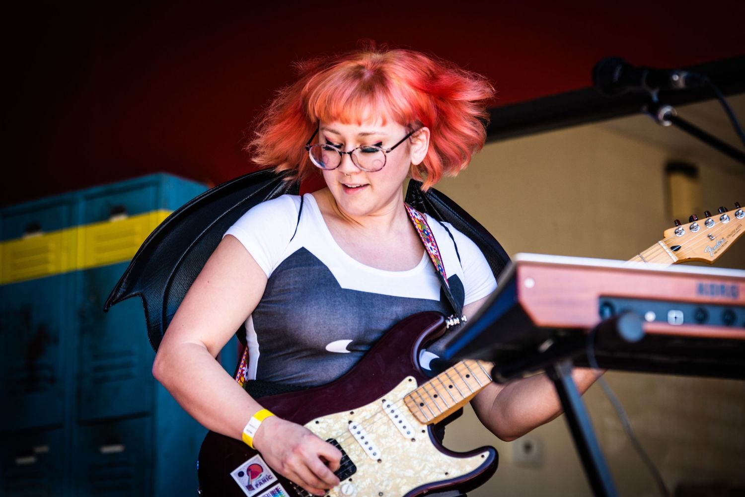 Guitarist performs energetically with pink hair and glasses under red stage lighting.