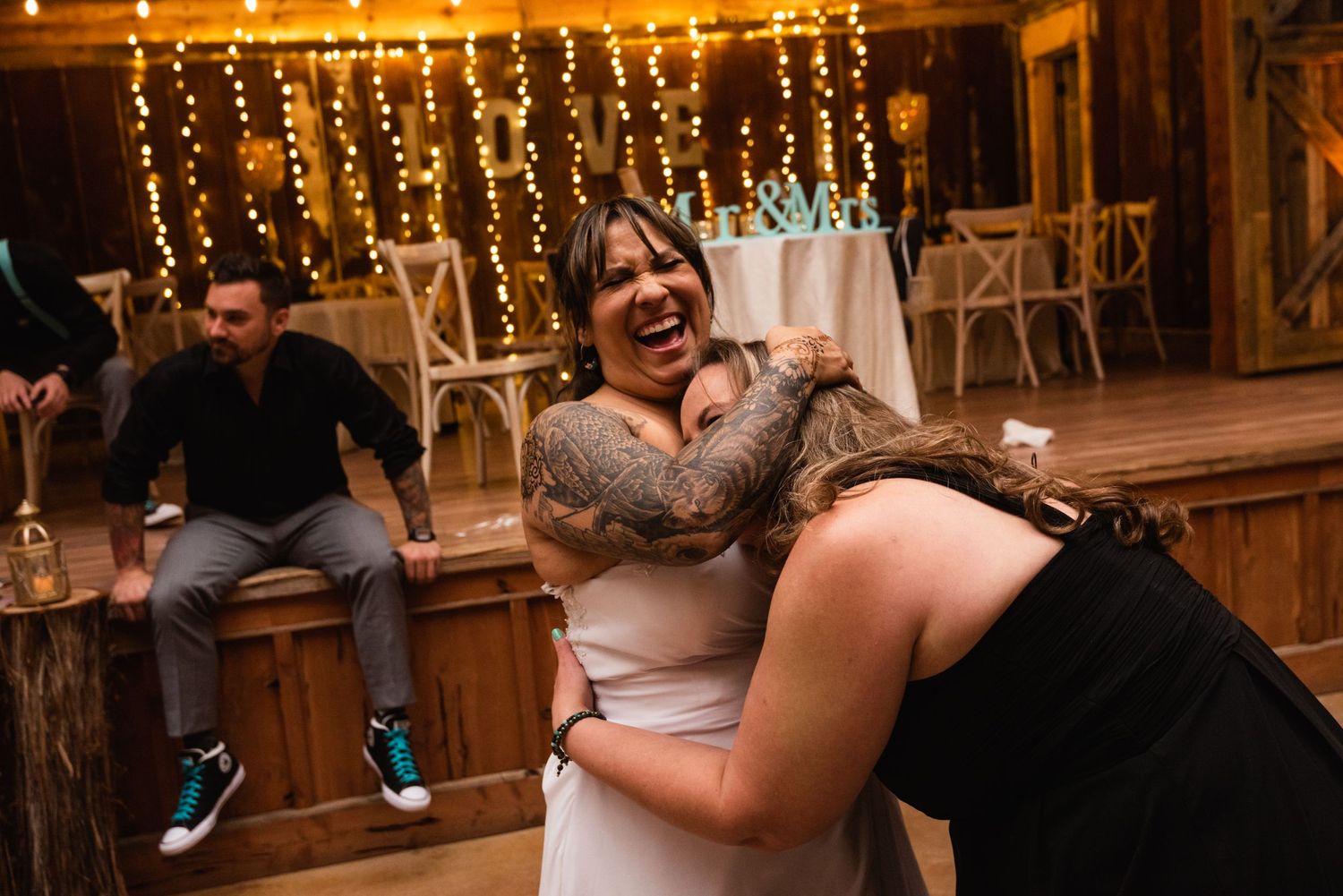 Wedding guests sharing a fun, candid moment under string lights at a rustic reception.