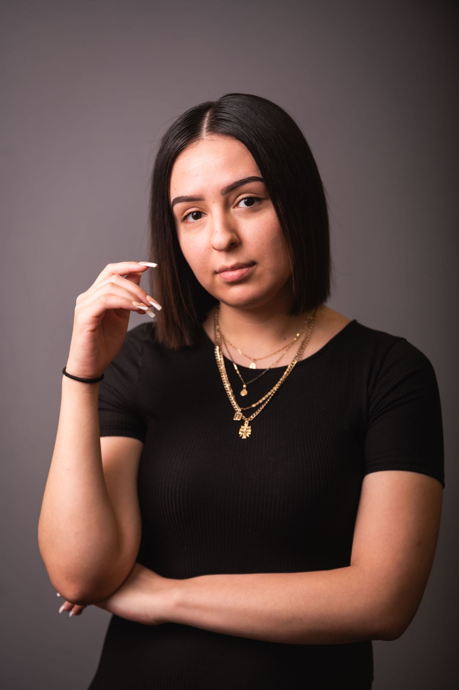 Artistic portrait of someone in black attire with layered gold necklaces against gray background.