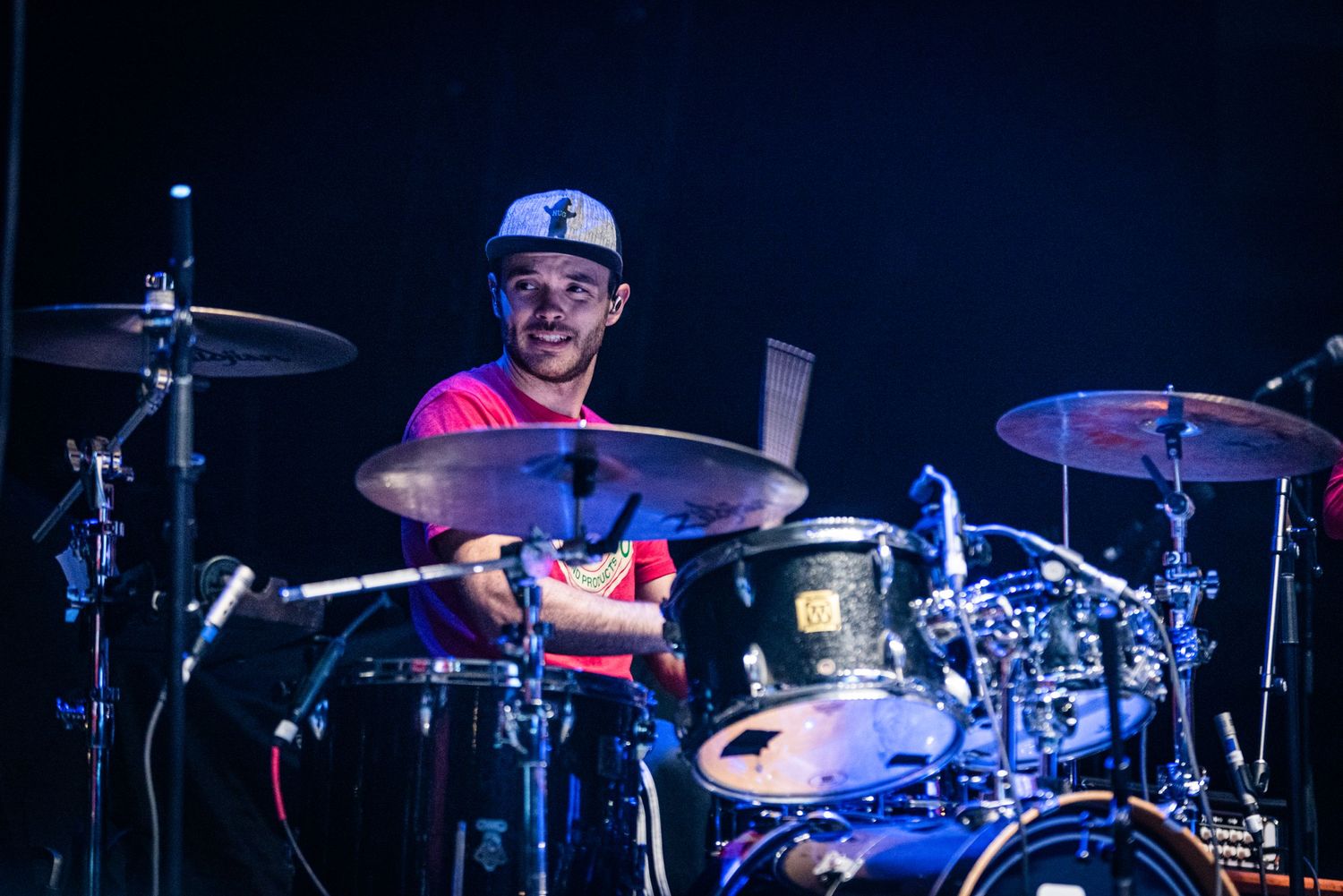 Drummer in blue baseball cap plays energetically on stage with dramatic lighting.