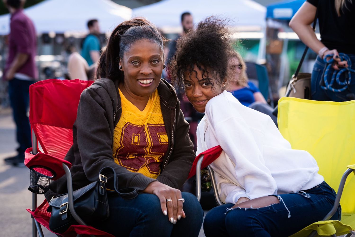 Two friends share a laugh while sitting in colorful camping chairs at outdoor event.