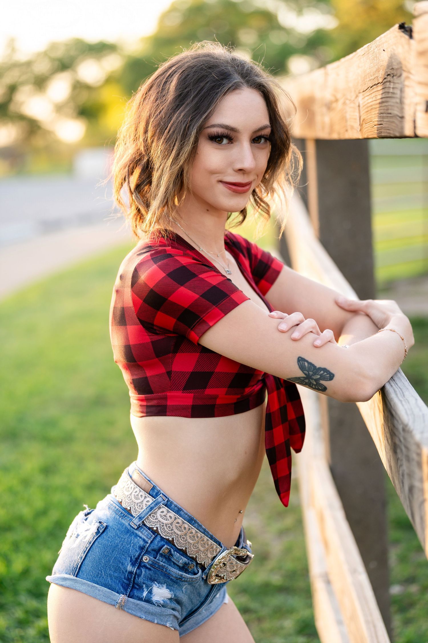Red plaid tied crop top and denim shorts outfit posed against rustic wooden fence.