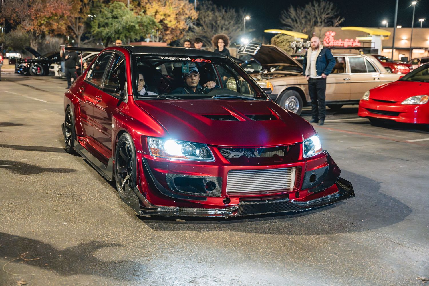 Modified red Mitsubishi Lancer Evolution with custom body kit and bright LED headlights at night.