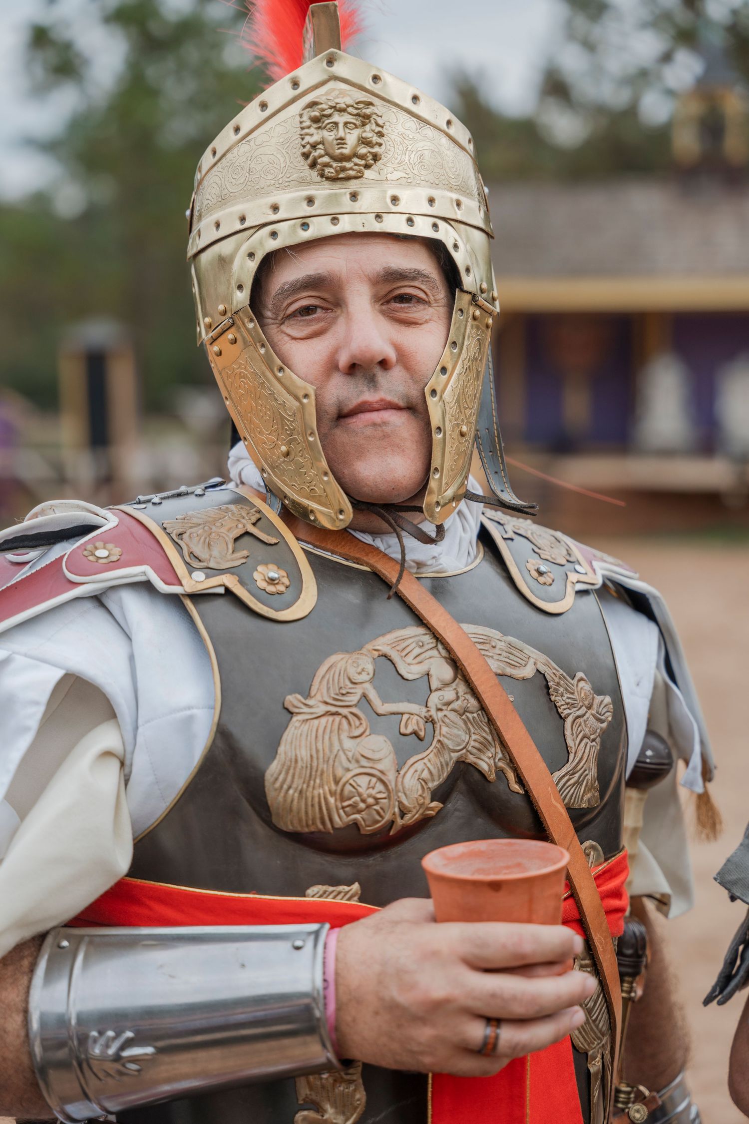 Performer in ornate Roman soldier armor and helmet holding clay cup at renaissance festival.