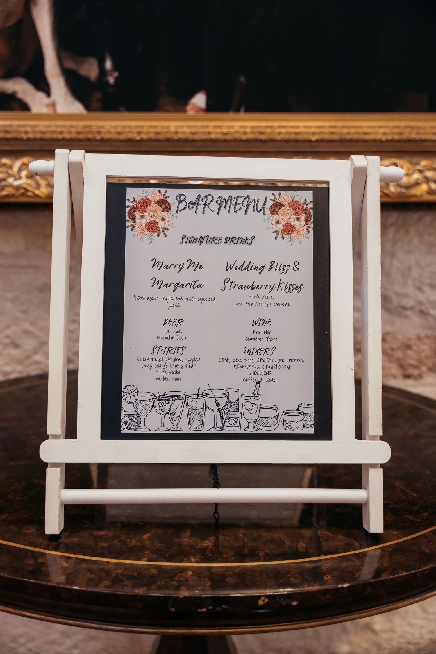 Wedding bar menu sign displayed on white easel with floral decorations.