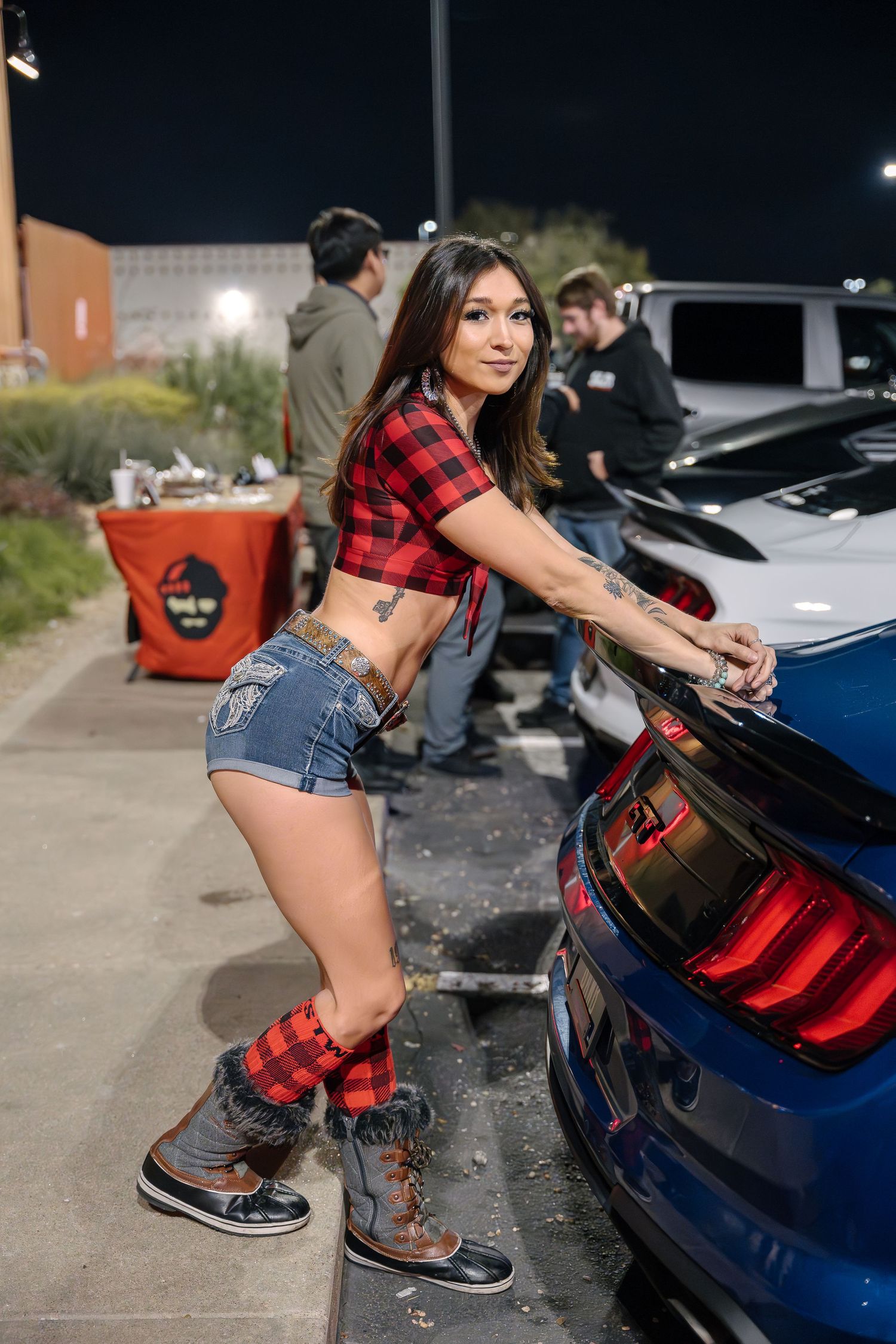 Red plaid crop top and denim shorts at a nighttime car meet next to a blue sports car.