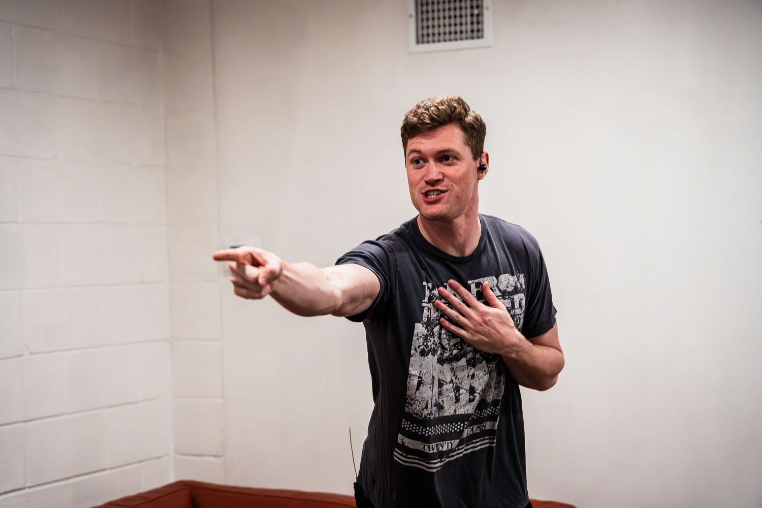 A performer gestures while standing in a white-walled dressing room.