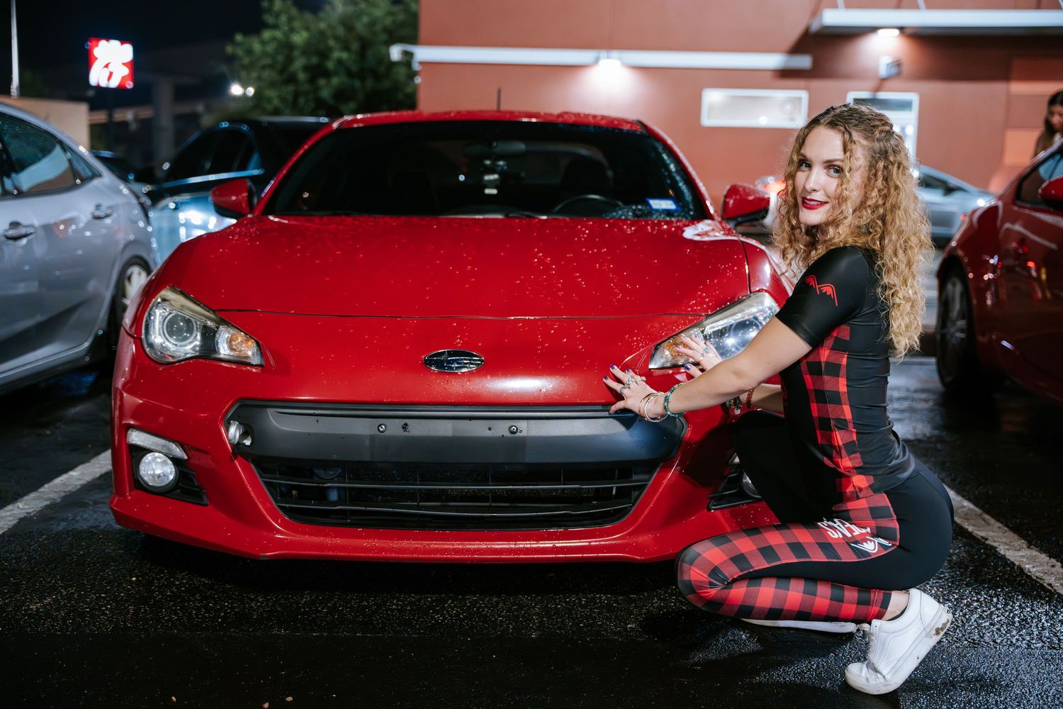 Crouching pose beside red Subaru BRZ at night car meet.