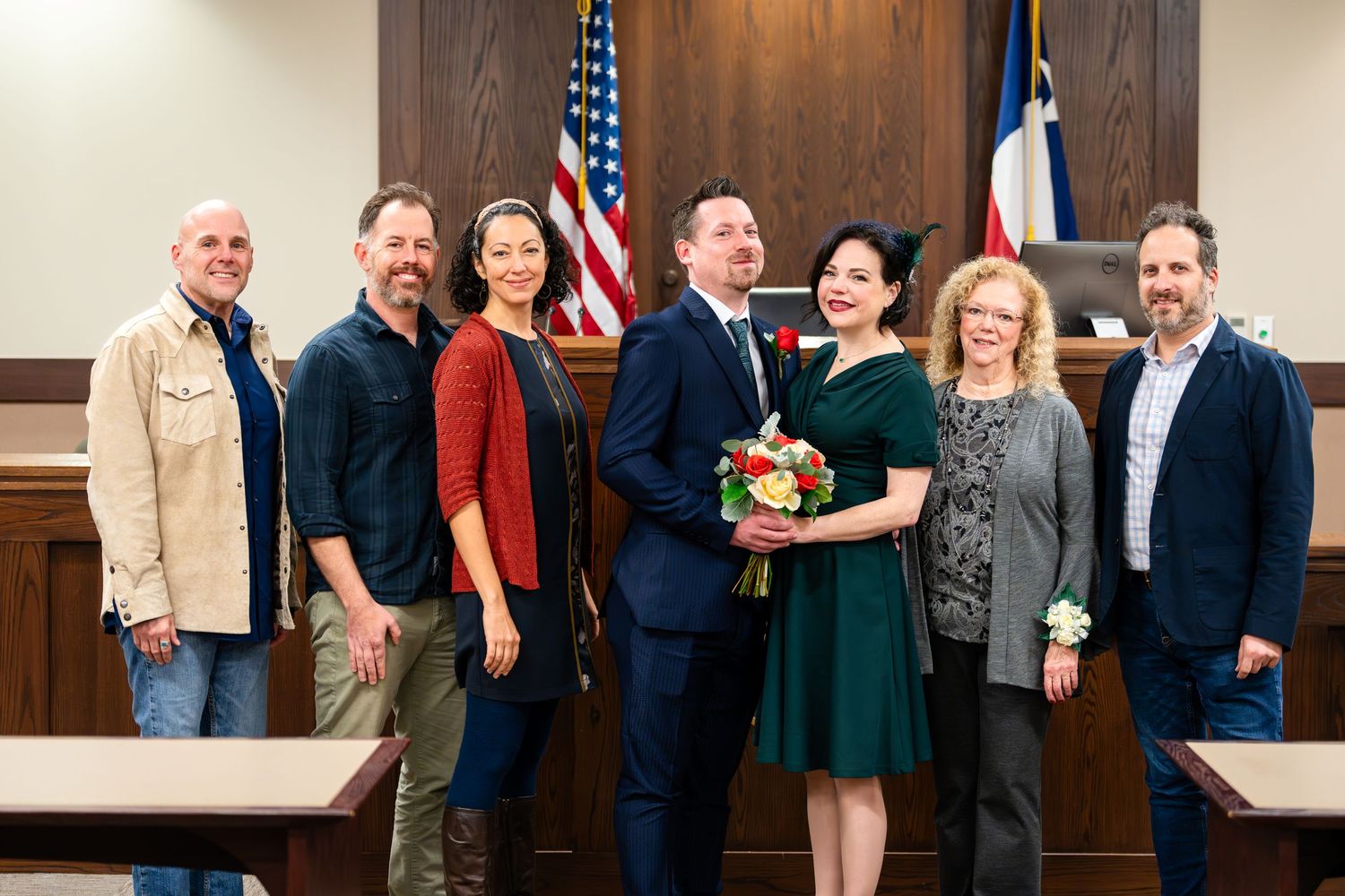 A group photo of wedding attendees gathered in a courthouse with American flags in the background.