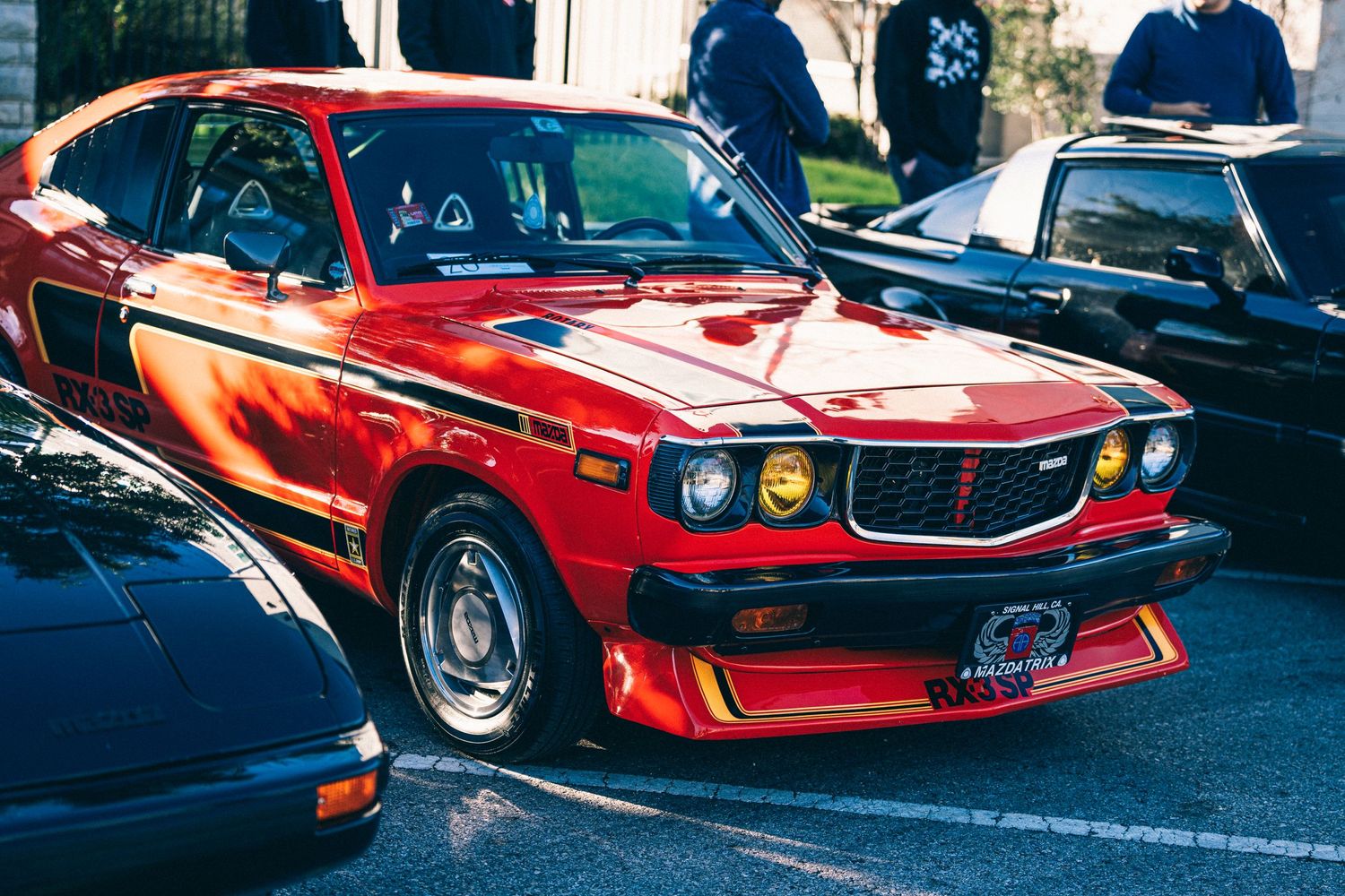Modified red Mazda RX-3 with wide body kit and racing stripes parked at a car show.