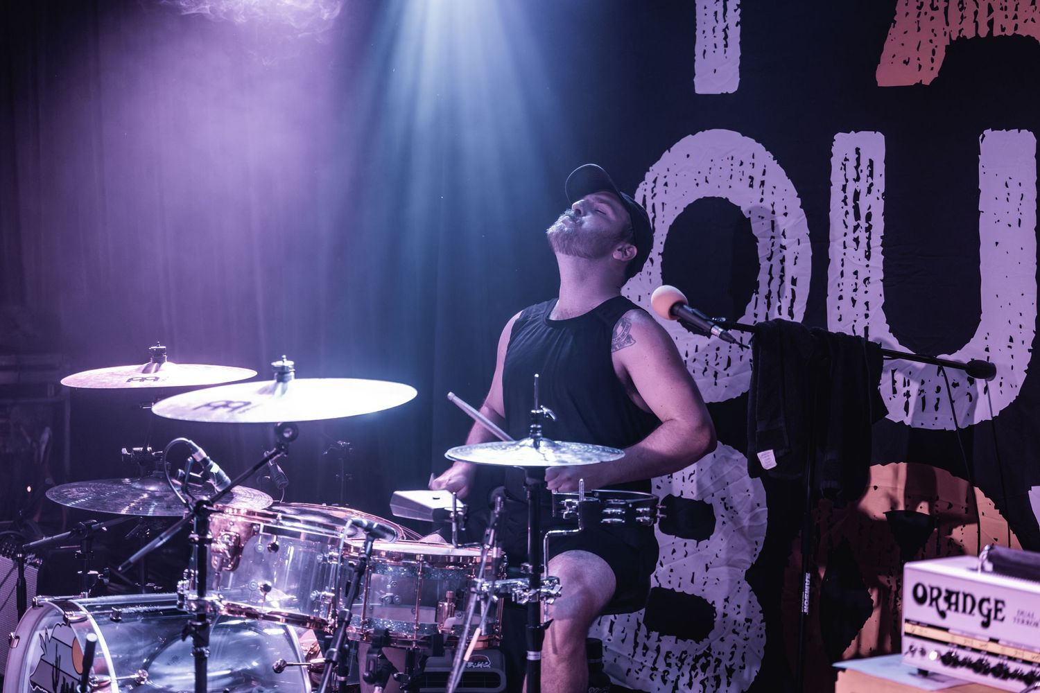 Drummer plays energetically on stage with purple and blue lighting effects.
