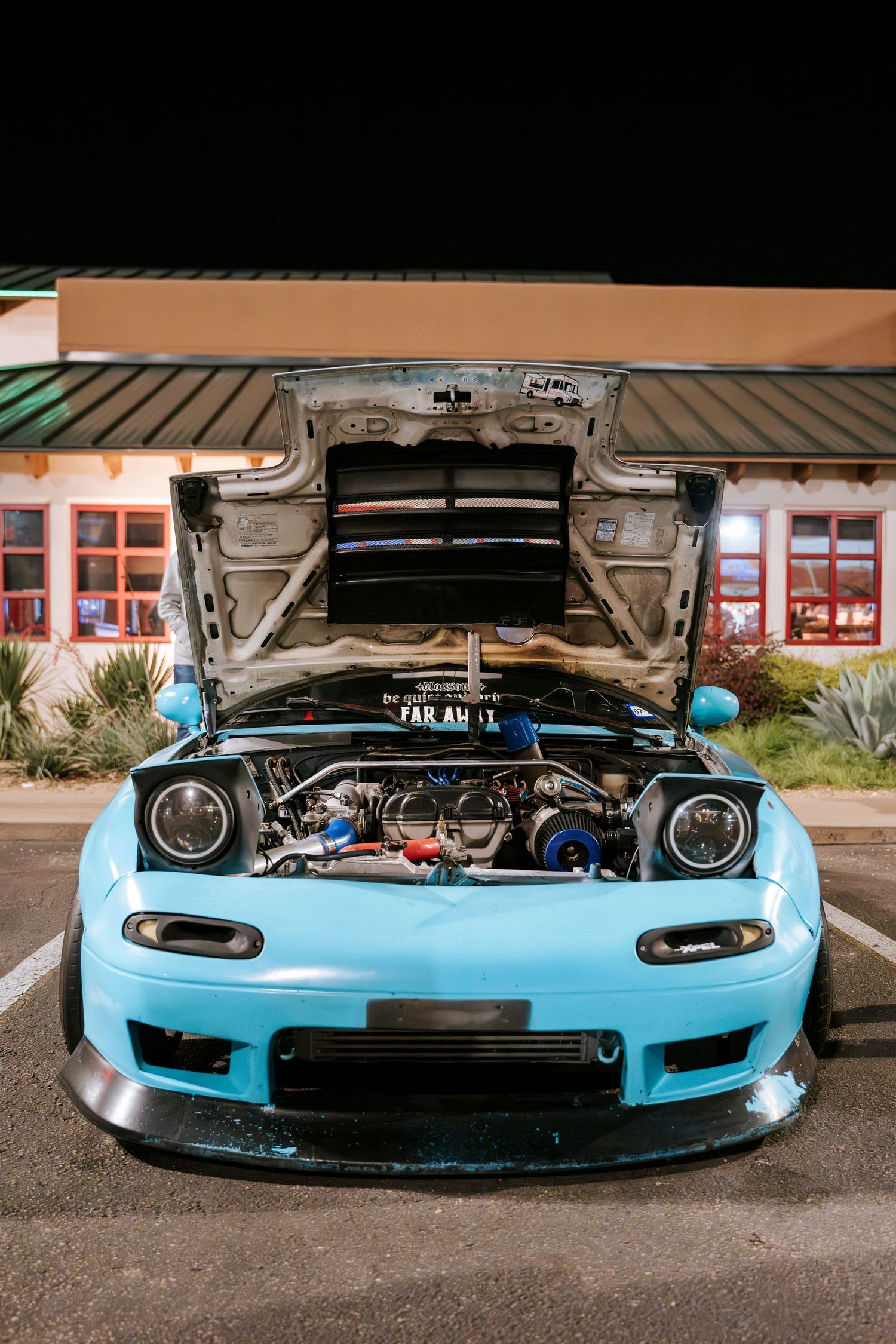 Light blue Mazda RX-7 with open hood showing modified engine bay at night.