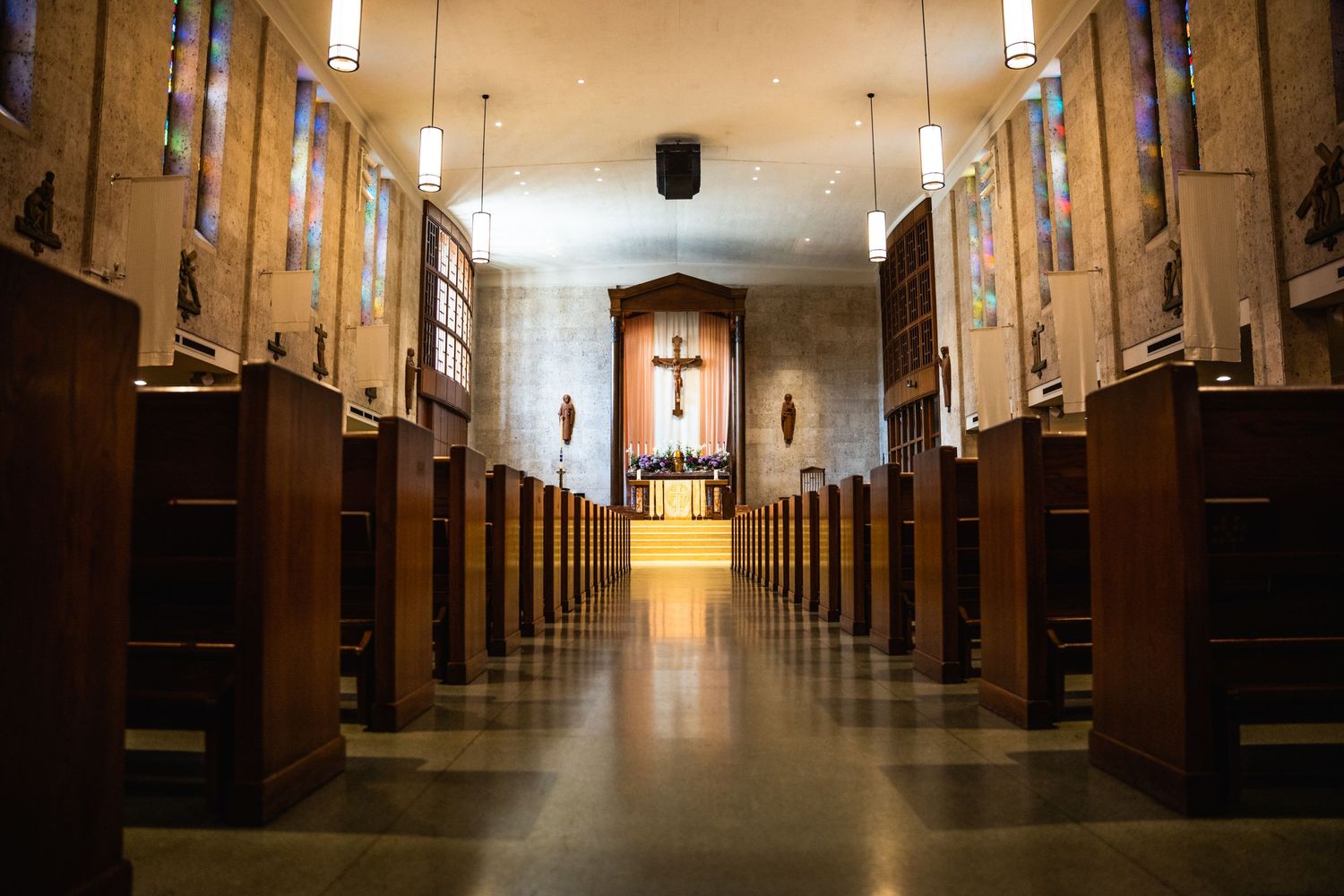 Empty church aisle leading to altar with wooden pews and hanging lights.