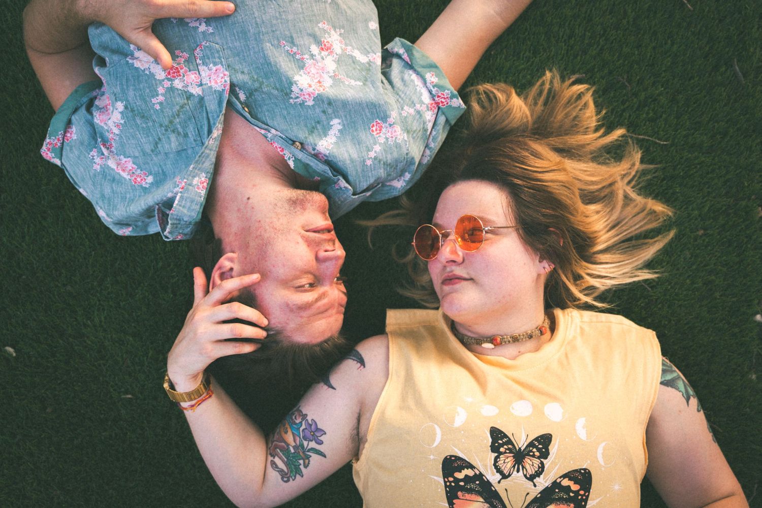 Two friends lying on grass taking creative perspective photo with vibrant clothing and sunglasses.