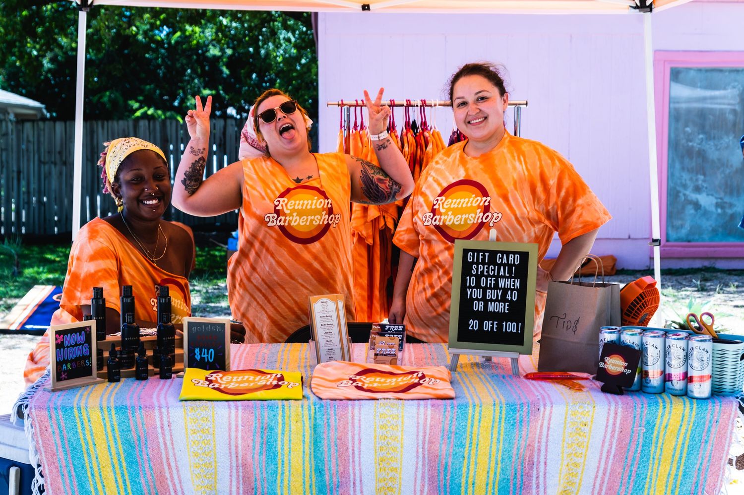 Vendors in orange shirts serve food and drinks at an outdoor booth with a colorful striped tablecloth.