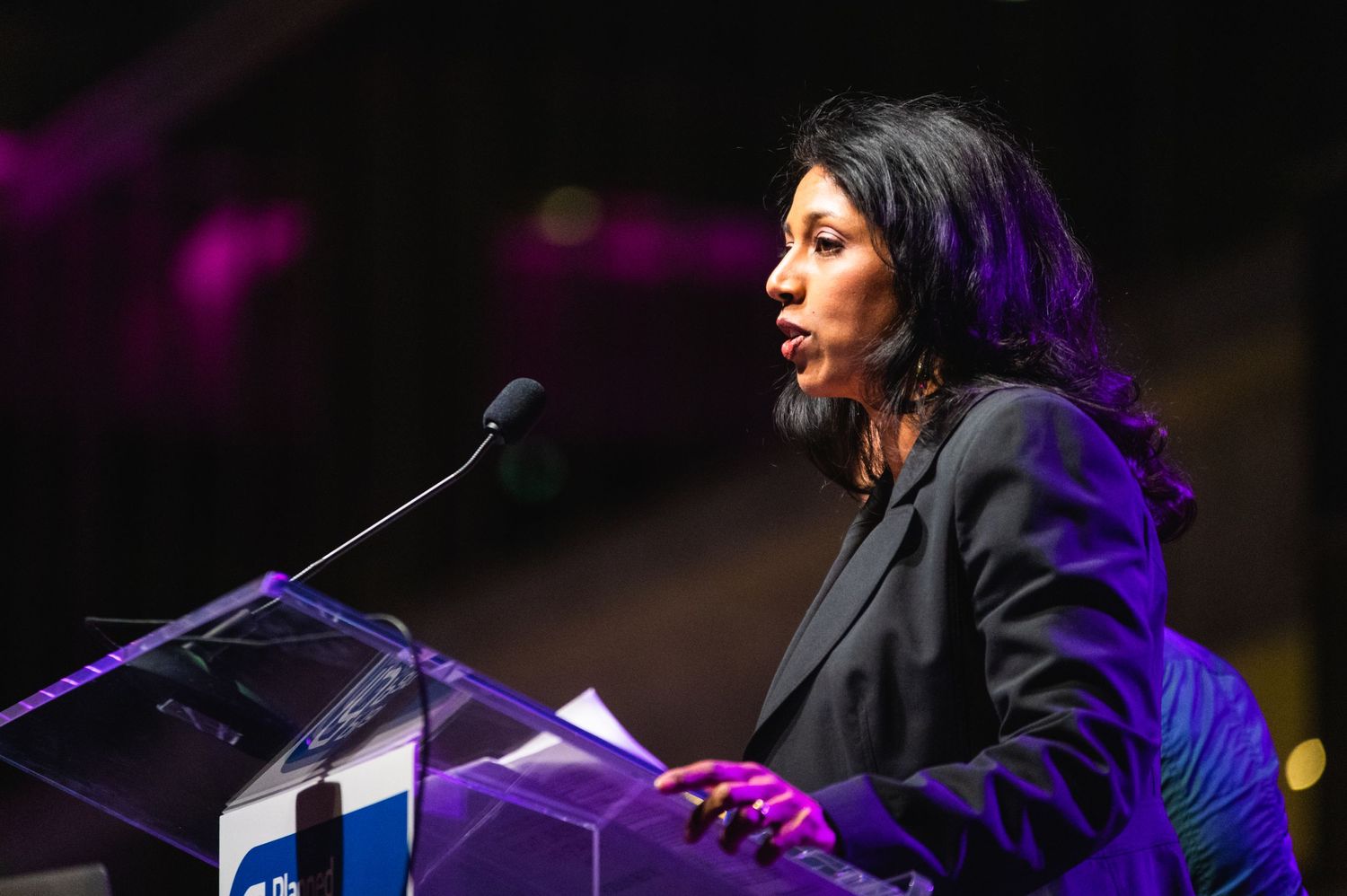 Speaker in dark blazer addresses audience from podium with purple stage lighting.