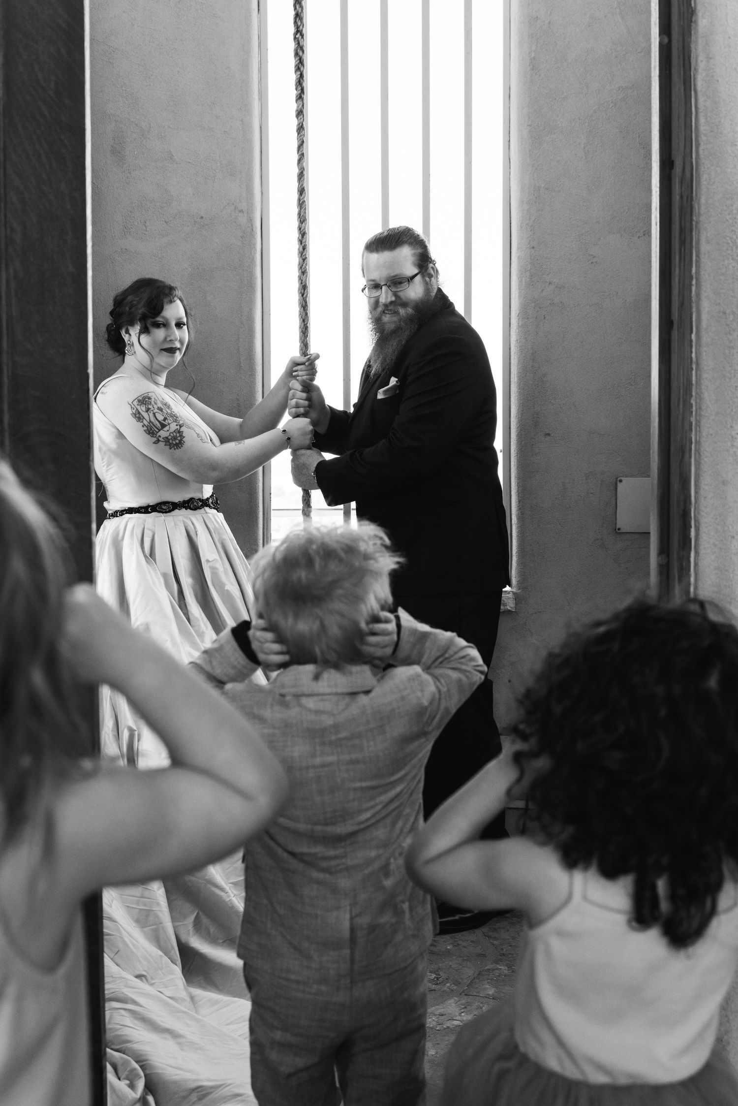 A playful black and white wedding photo showing a couple posing with a rope by a window while guests look on.