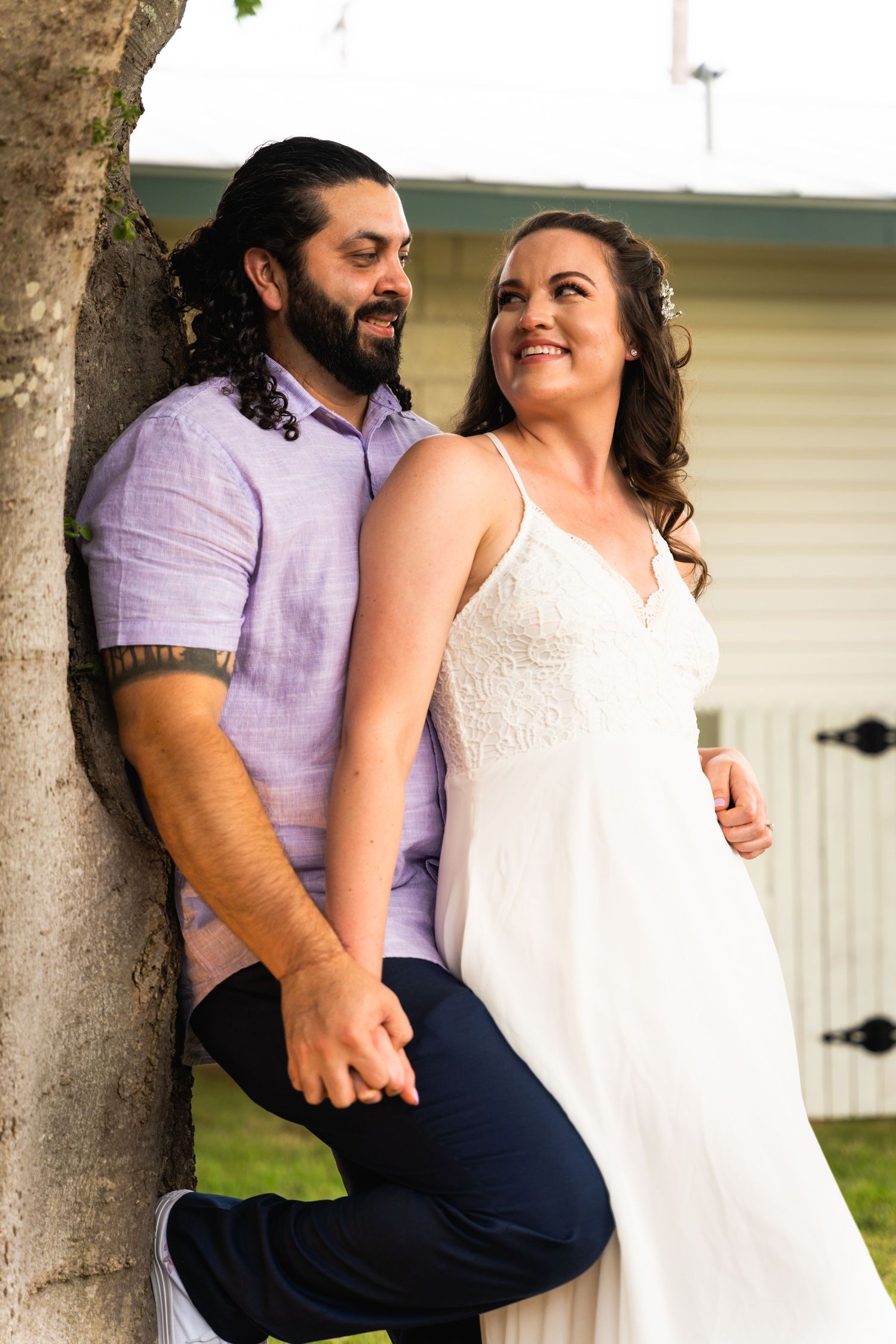 A couple shares a candid moment together by a tree in casual wedding attire.