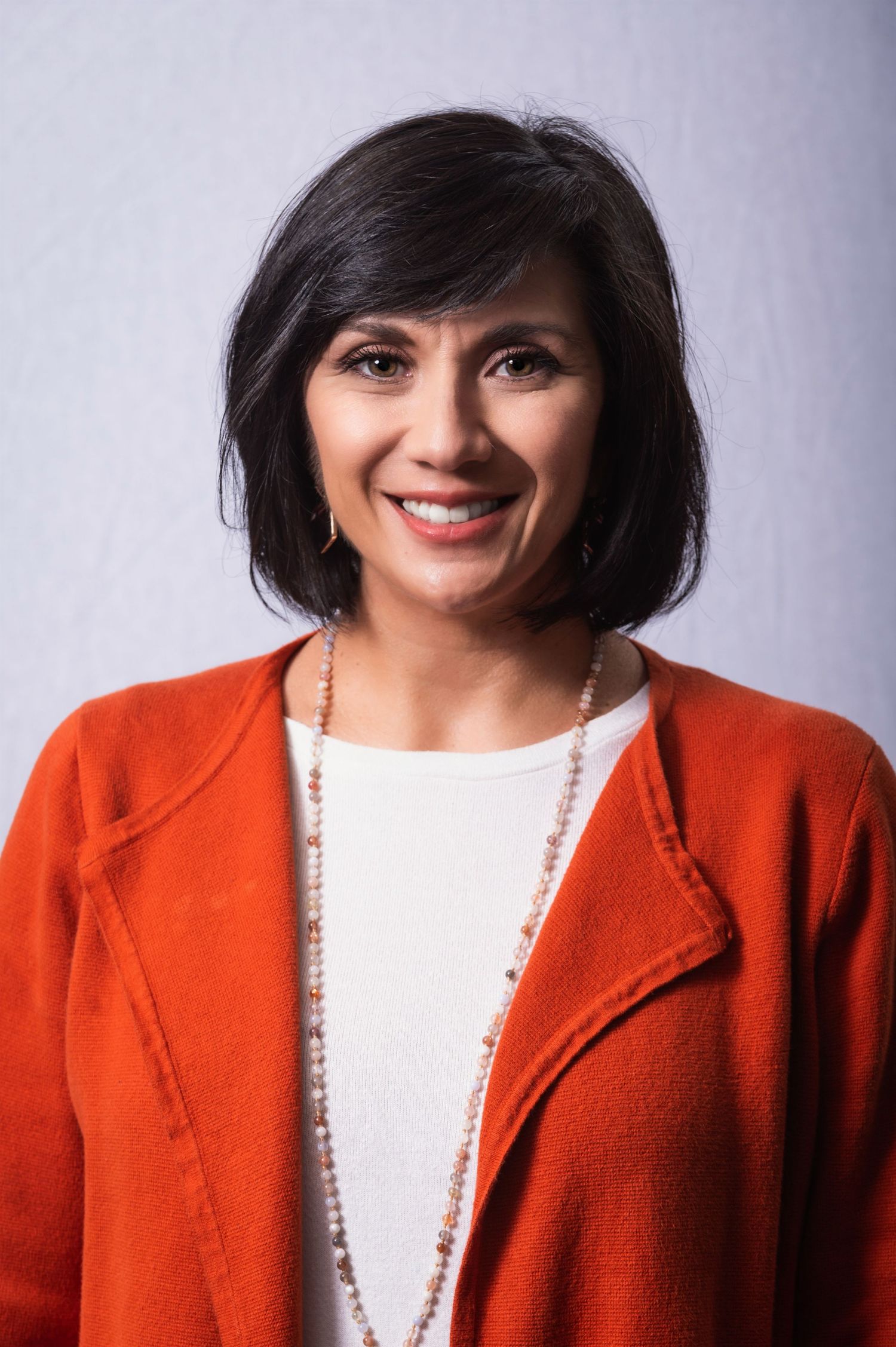 Studio portrait of someone in an orange cardigan and white top with a beaded necklace against gray background.