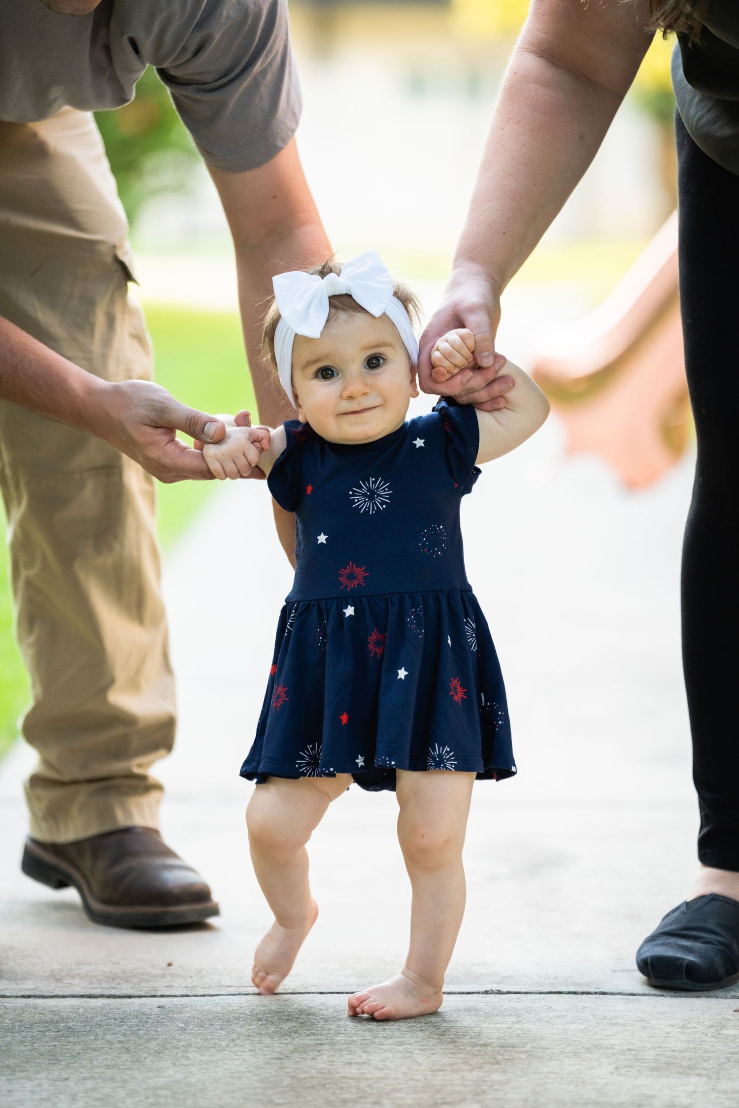 A toddler in a navy dress takes first steps while supported by adult hands.