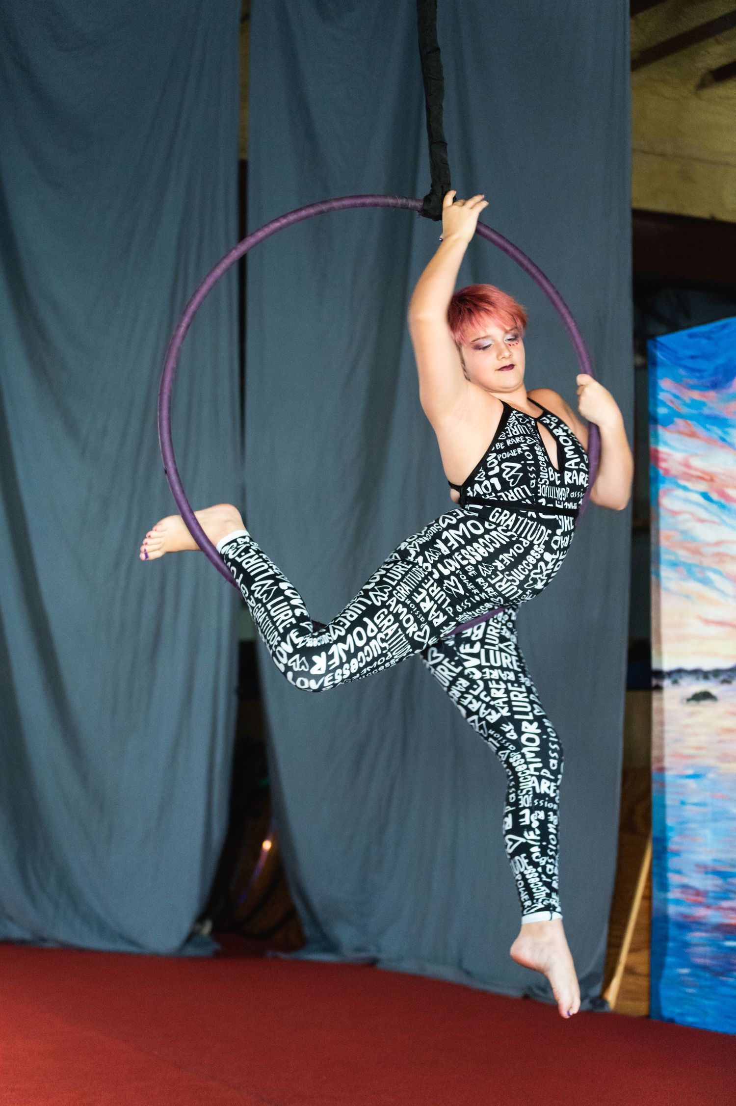 Aerialist in patterned outfit performs dynamic pose on suspended purple hoop.