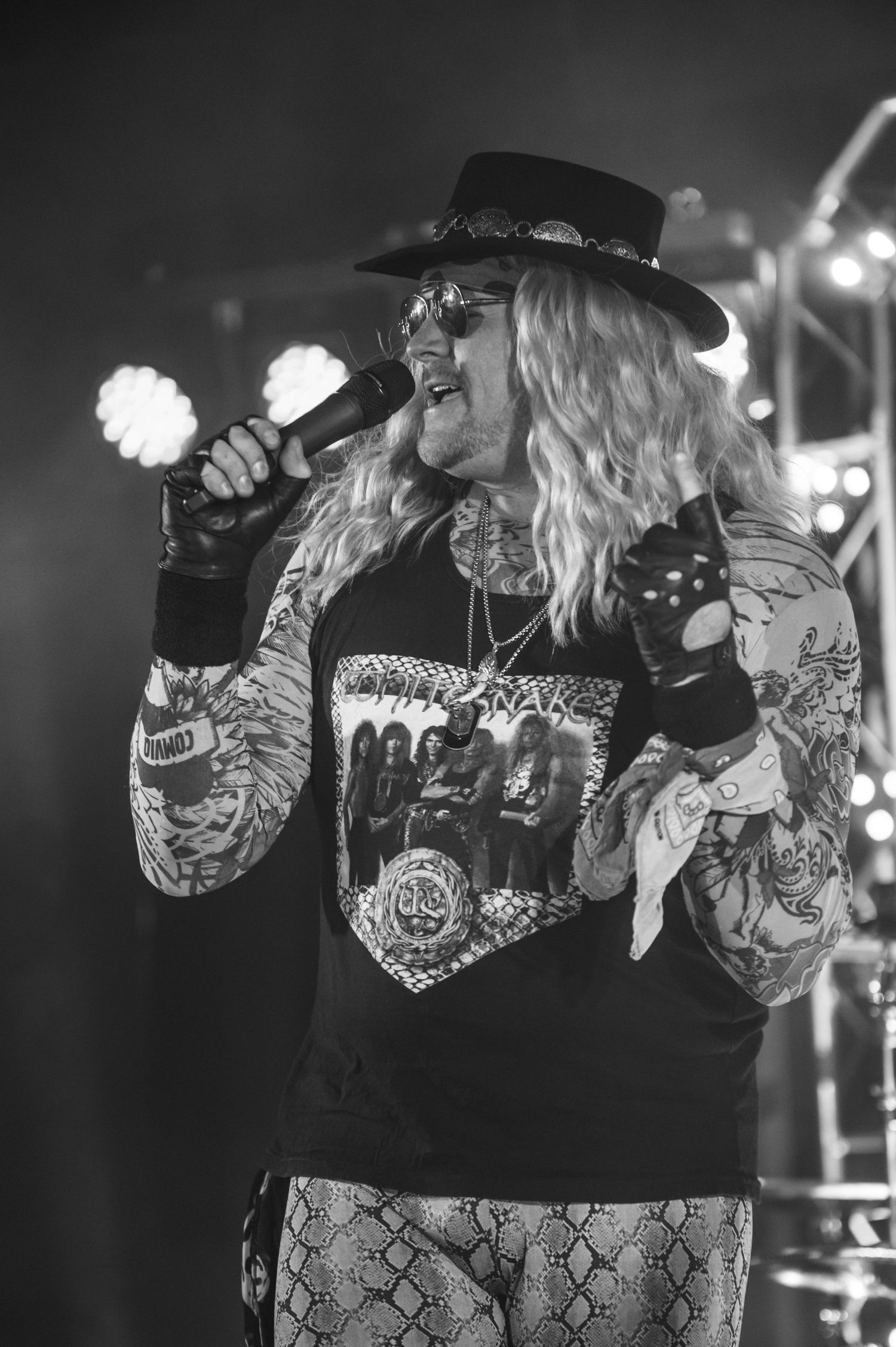 Rock singer performs on stage wearing a hat, band t-shirt and snakeskin pants under concert lighting.